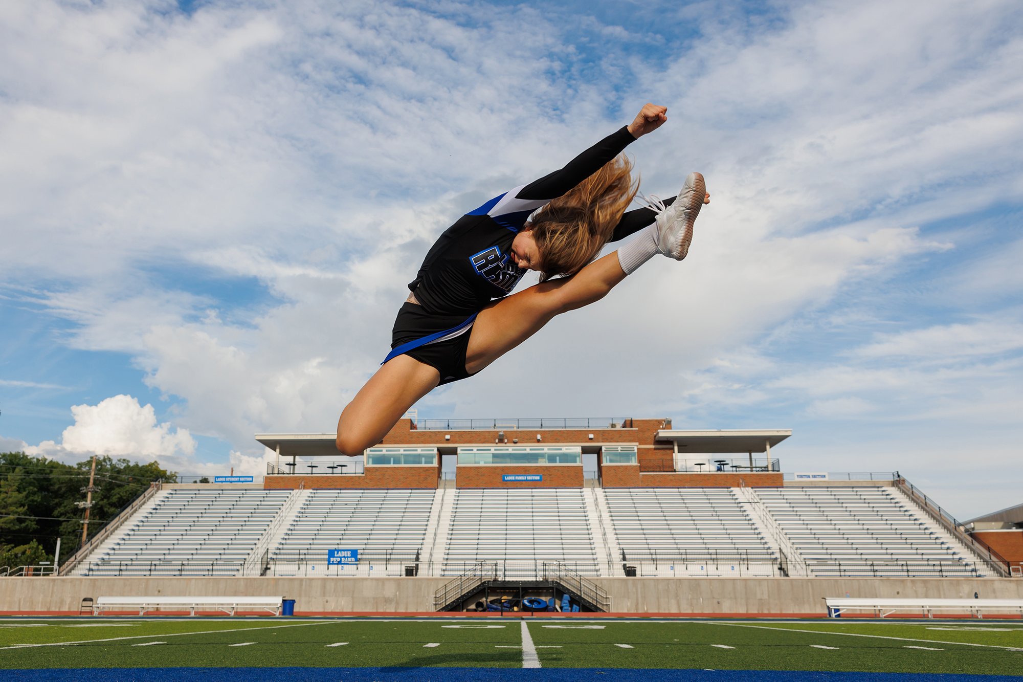 Female athlete mid-air performing a high jump over a football field with empty stadium seats and blue sky in the background.