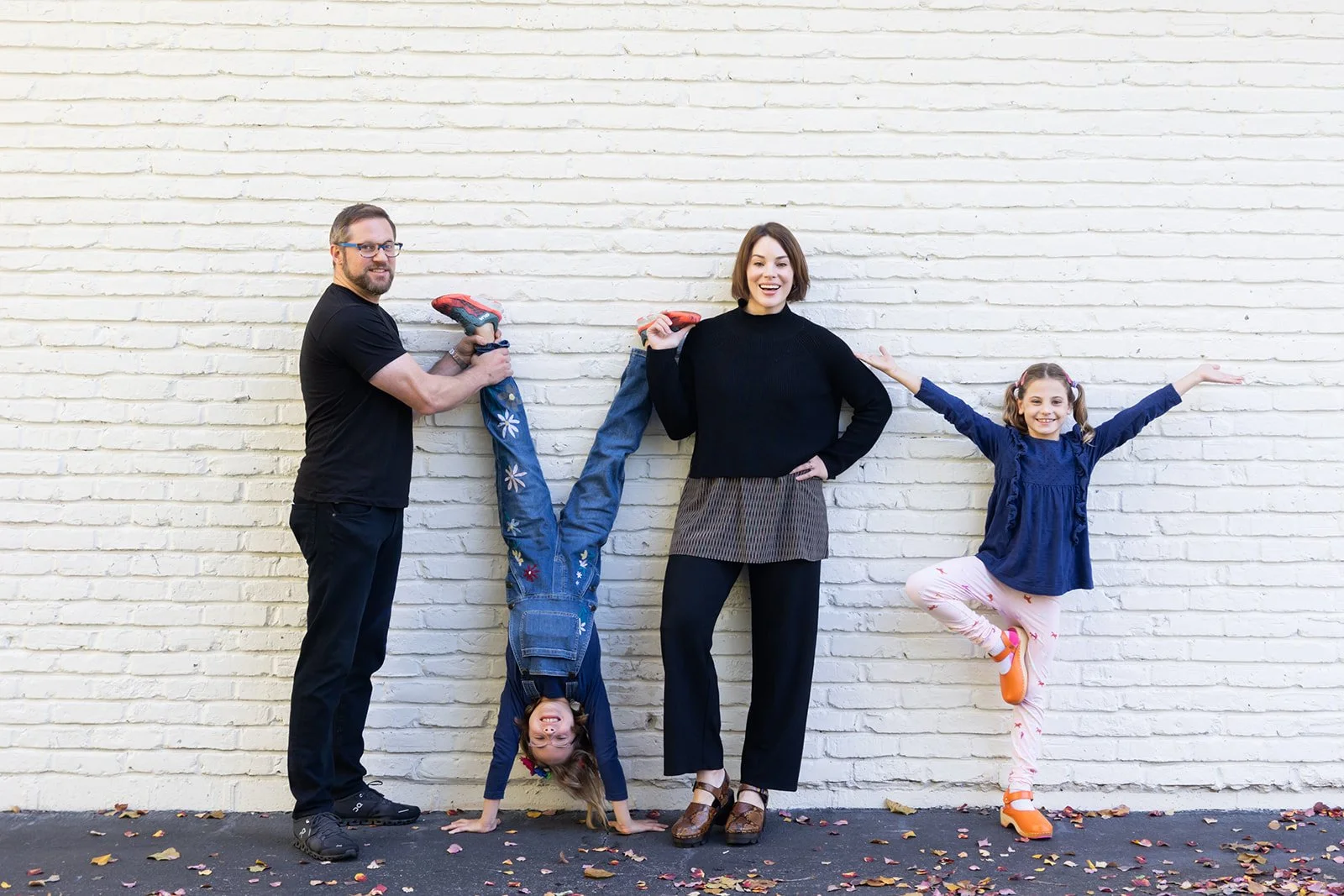 A family of four standing against a white brick wall, with a man and a woman holding up two young girls. One girl is doing a handstand, and the other is standing on one leg with arms outstretched.