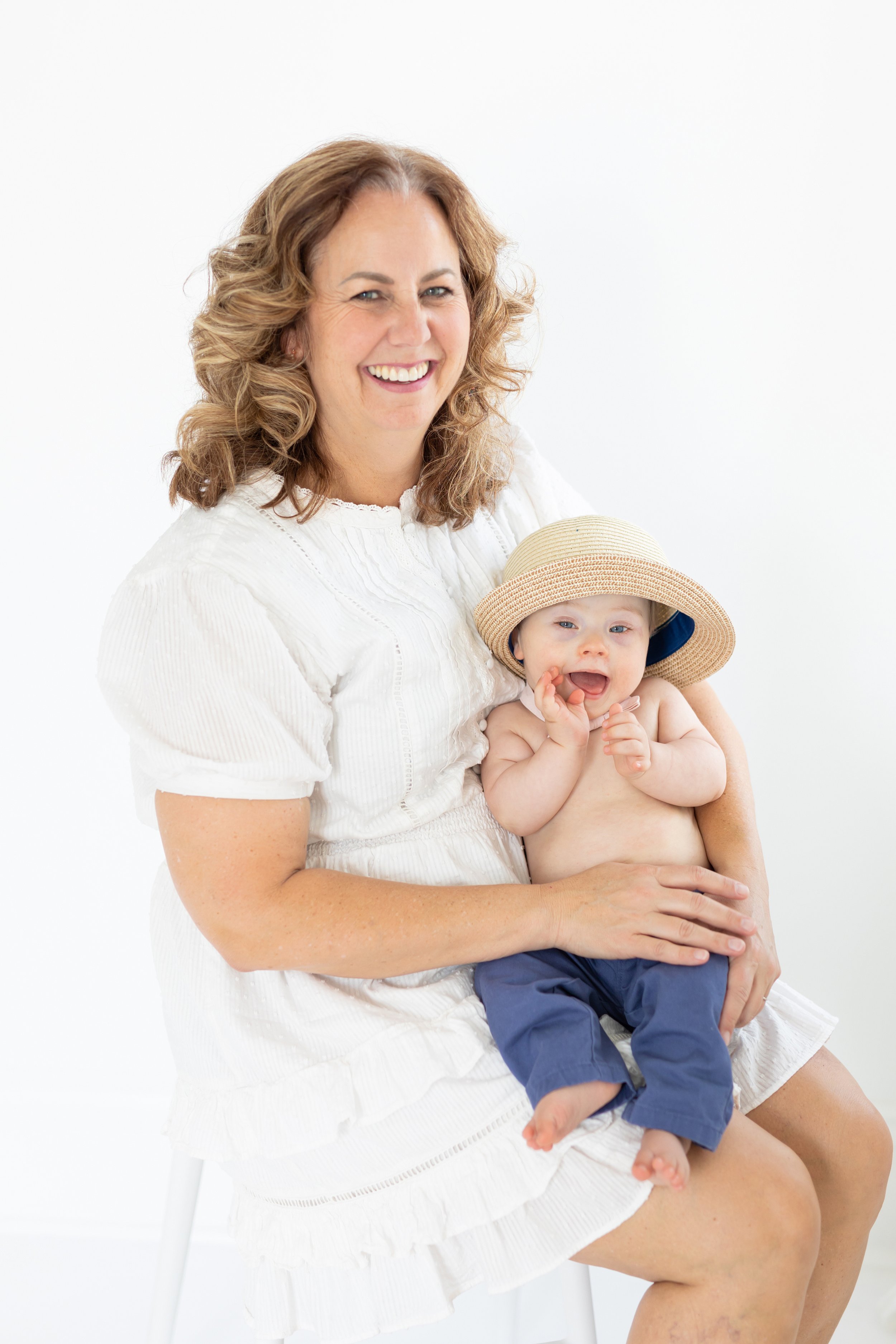 A woman holding a toddler boy wearing a sunhat, both sitting on a white stool against a white background, smiling and enjoying each other's company.