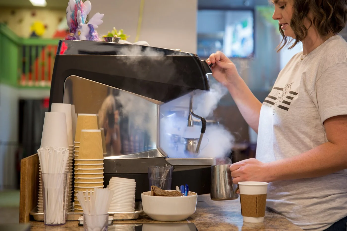 Barista preparing coffee with steam emerging from the espresso machine at a coffee shop, with paper cups, stir sticks, and a plastic cup on the counter.
