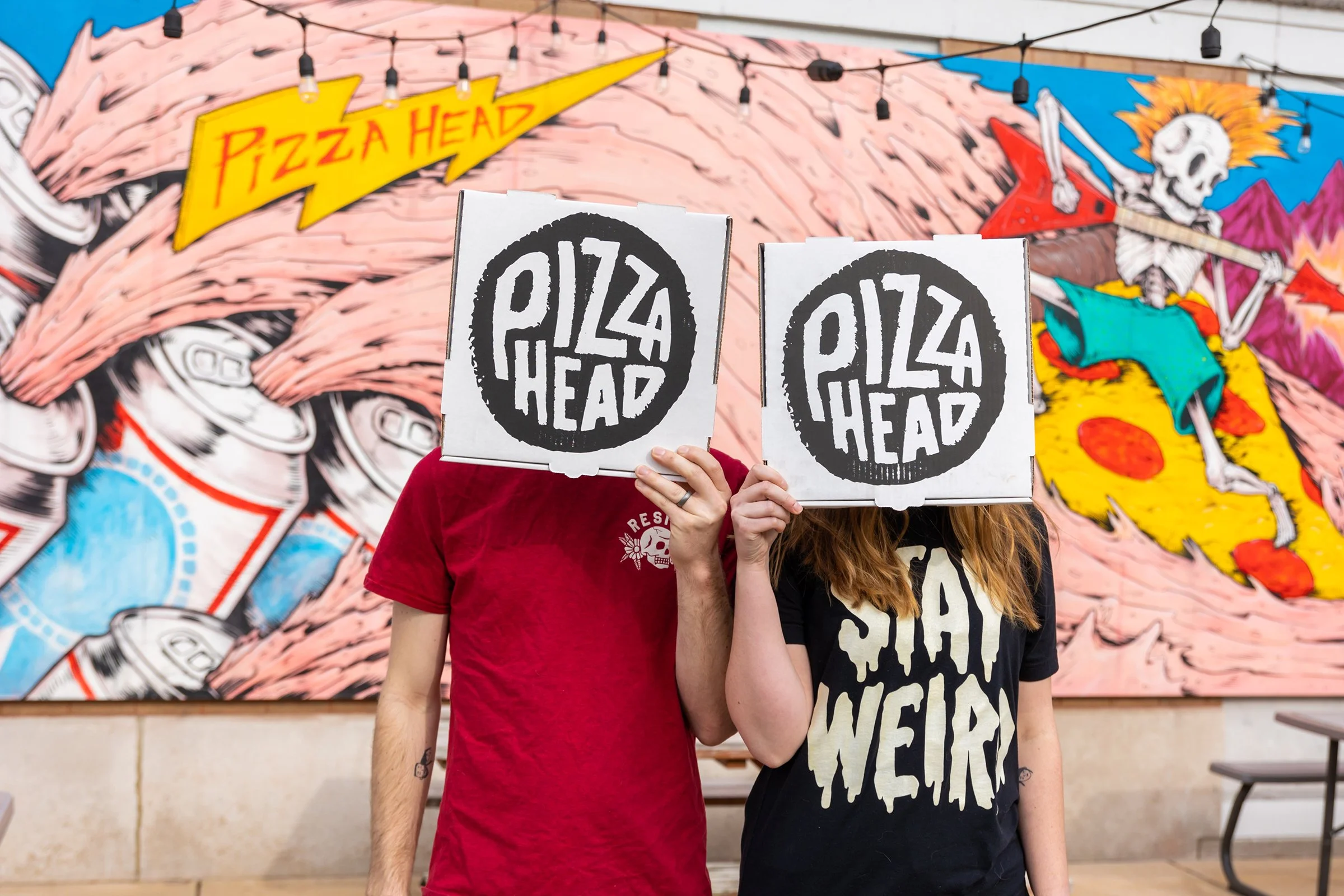 Two people holding up boxes with the 'Pizza Head' logo covering their faces, standing in front of a colorful mural with a cartoonish skeleton playing guitar dressed in a cape and a tutu, with a banner that says 'Pizza Head' and various cartoon illustrations.
