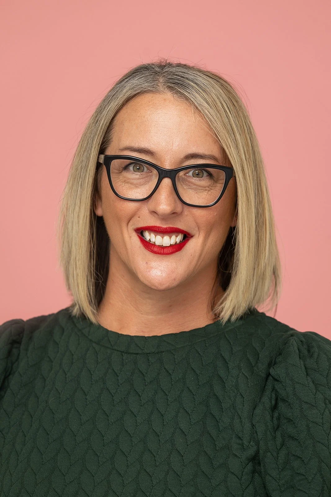 Smiling woman with shoulder-length blonde hair, black glasses, red lipstick, wearing a dark green textured top, against a pink background.