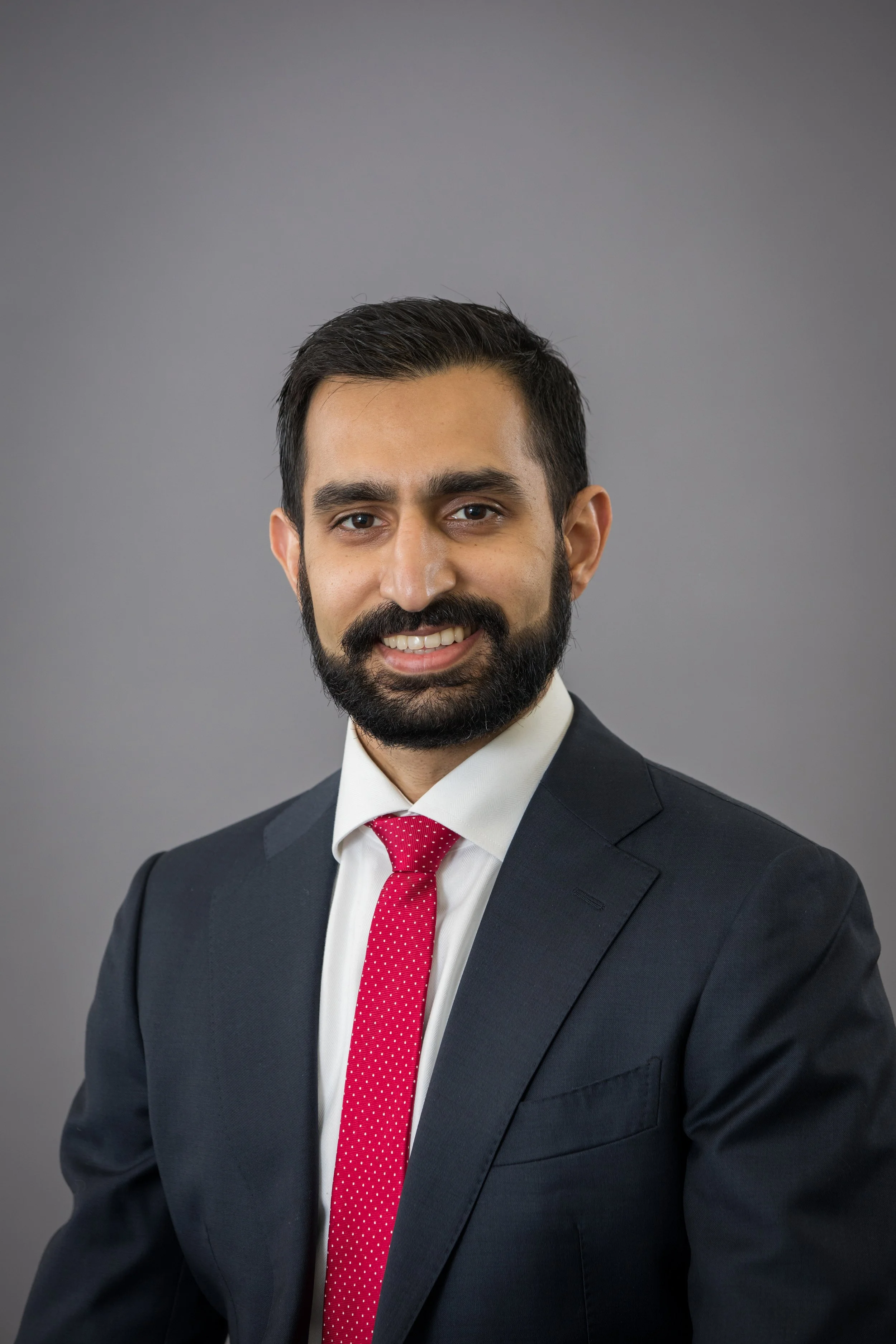 Professional portrait of a man with dark hair, a full beard, wearing a black suit, white shirt, and red polka dot tie, smiling against a gray background.