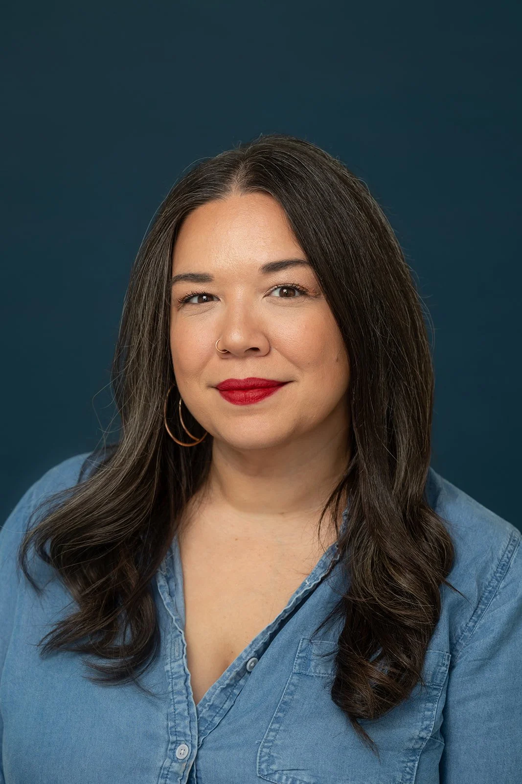 A woman with dark brown hair styled in loose waves, wearing a denim button-up shirt, red lipstick, small hoop earrings, and a nose ring, posed against a dark blue background.
