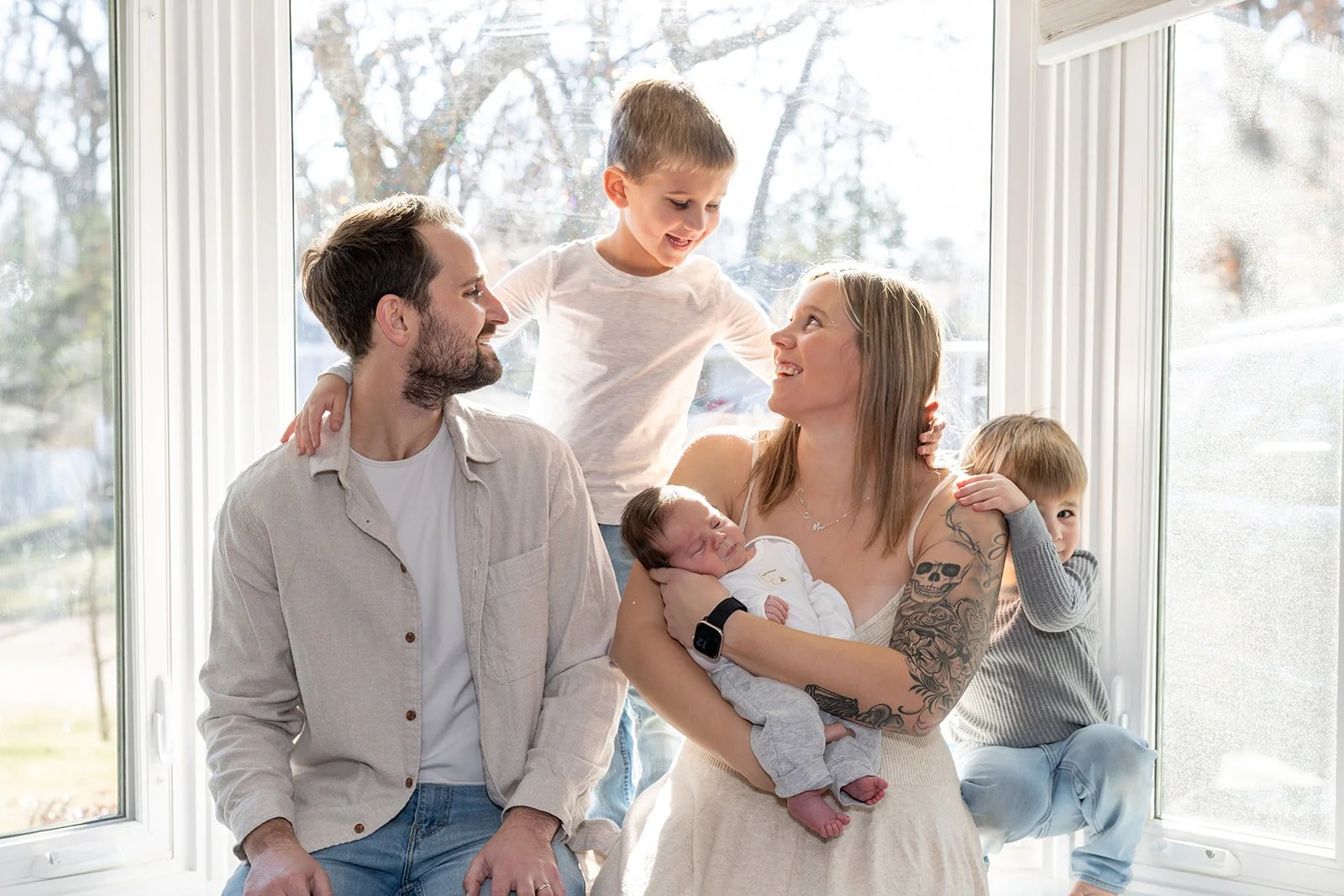 A family of six posing together in front of a large window, with a woman holding a newborn baby and a young boy sitting on the window sill, while a man and a young girl stand beside them, all smiling and happy.