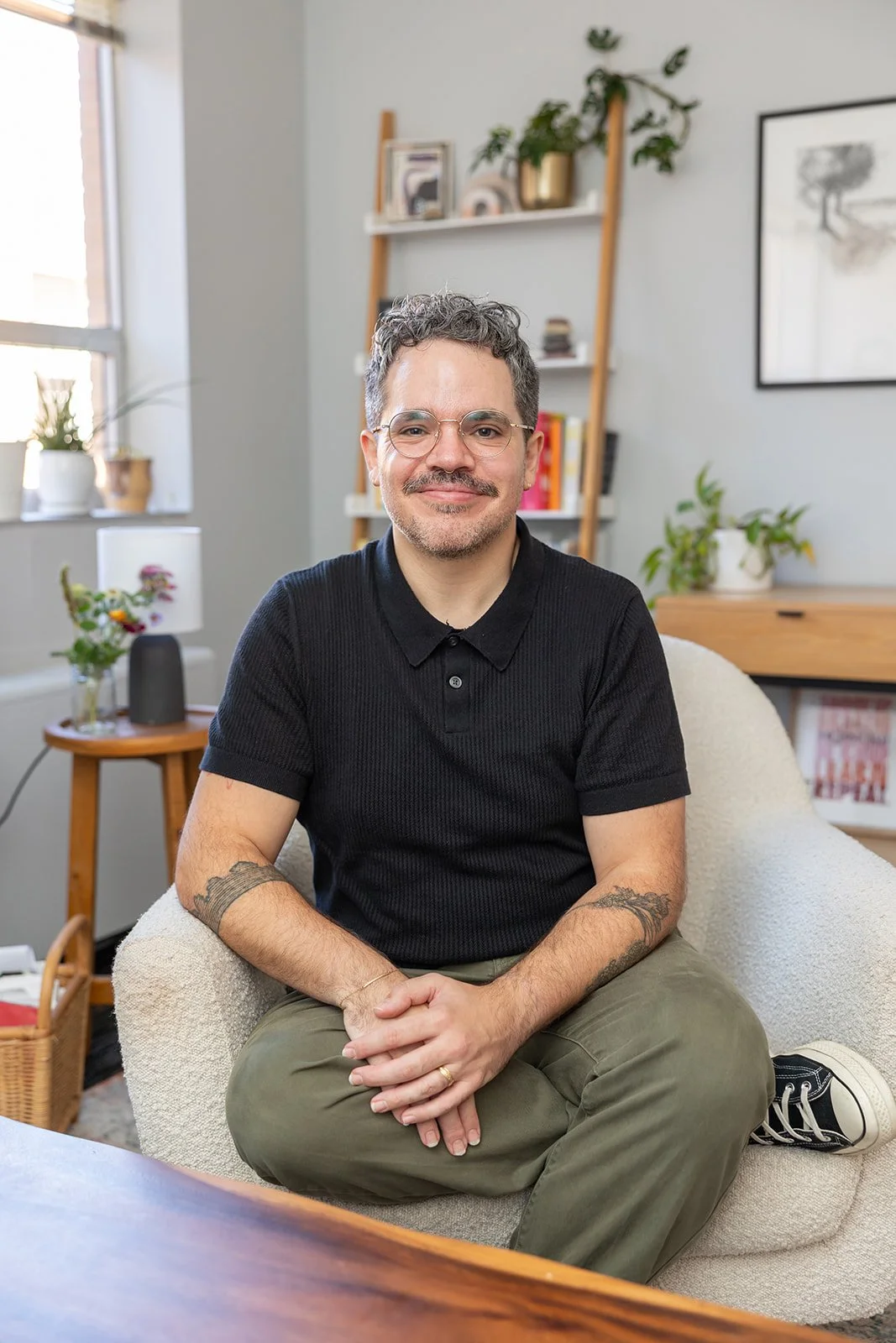 A man with curly hair, glasses, and tattoos, sitting cross-legged on a cream-colored armchair in a well-lit room with shelves, plants, and artwork in the background.
