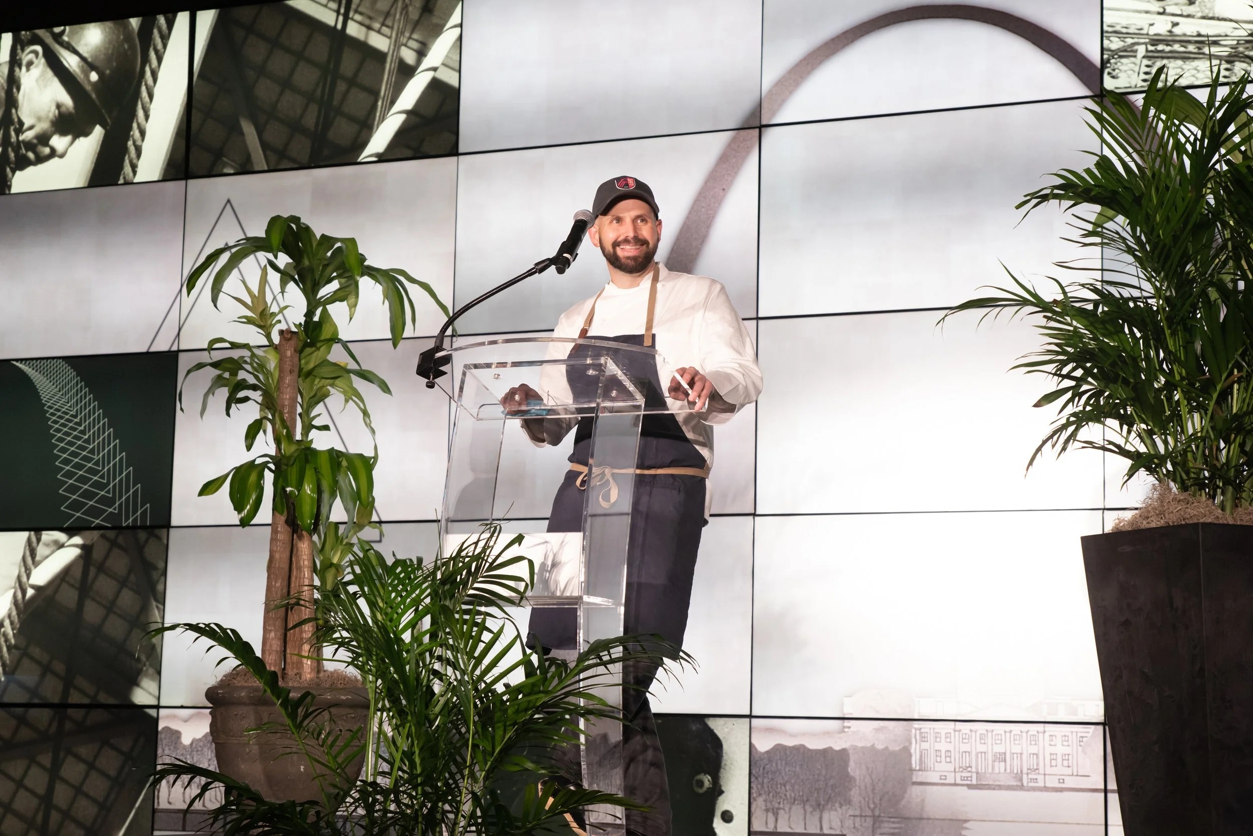 A man giving a speech at a podium with a microphone, surrounded by green plants, with multiple large screens displaying black and white images of a person and architecture backgrounds.