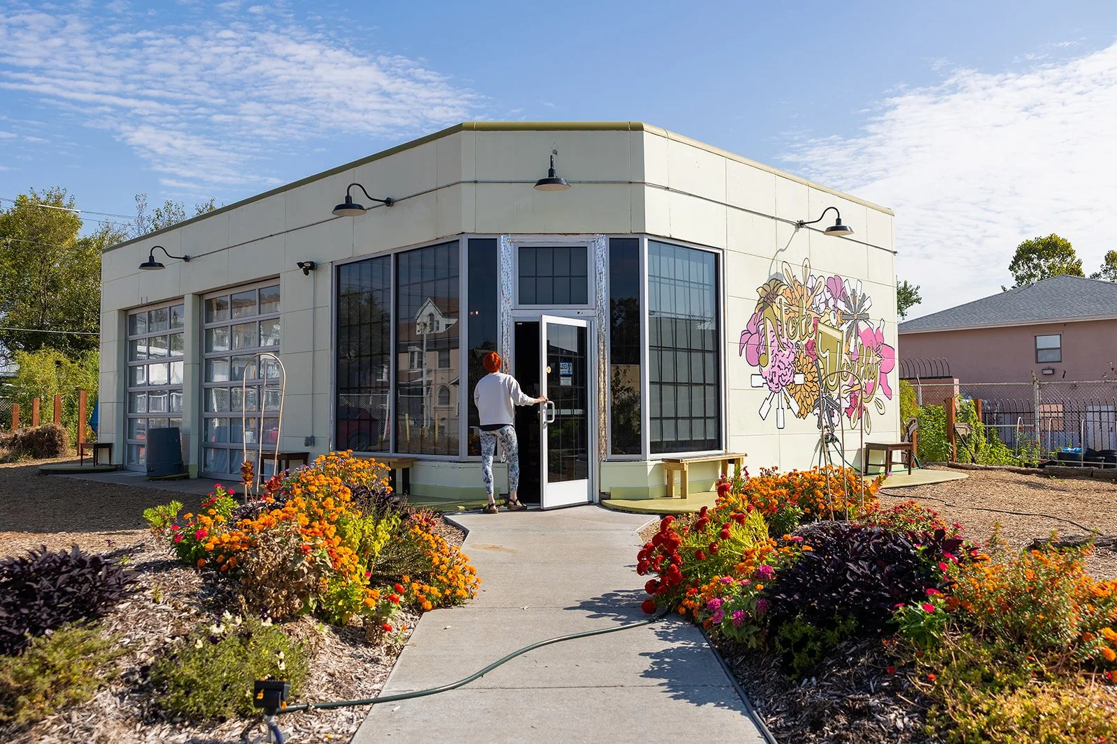 A small modern building with large windows and a colorful mural on the side, surrounded by flower beds and a sidewalk, with a person entering the door on a sunny day.