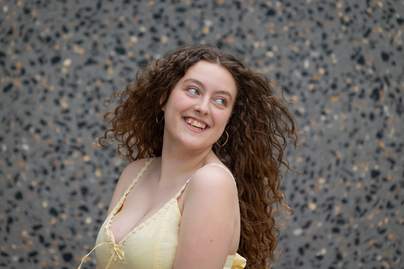 A young woman with curly brown hair wearing a yellow spaghetti strap top, looking to her left and smiling, standing in front of a textured, multicolored stone wall.