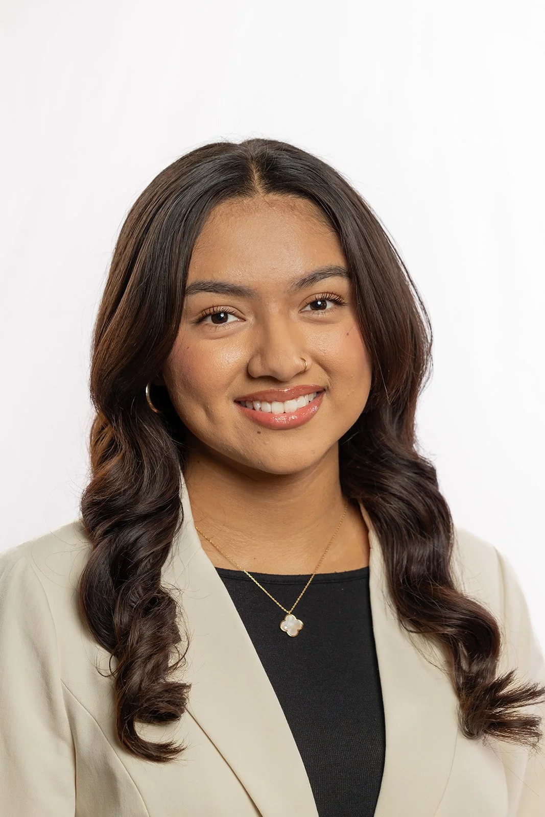Portrait of a young woman with wavy dark hair, wearing a light-colored blazer, black top, gold necklace with a four-leaf clover pendant, and small hoop earrings, smiling against a white background.