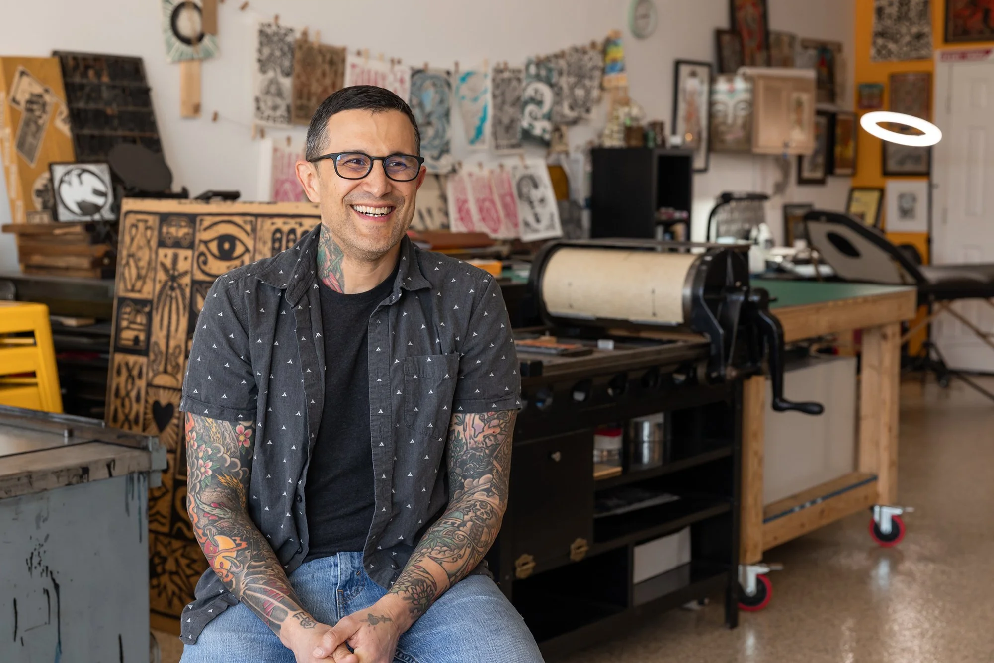 Man with tattoos wearing glasses, black shirt, and patterned short-sleeve shirt smiling in an art studio filled with printmaking equipment and artwork.