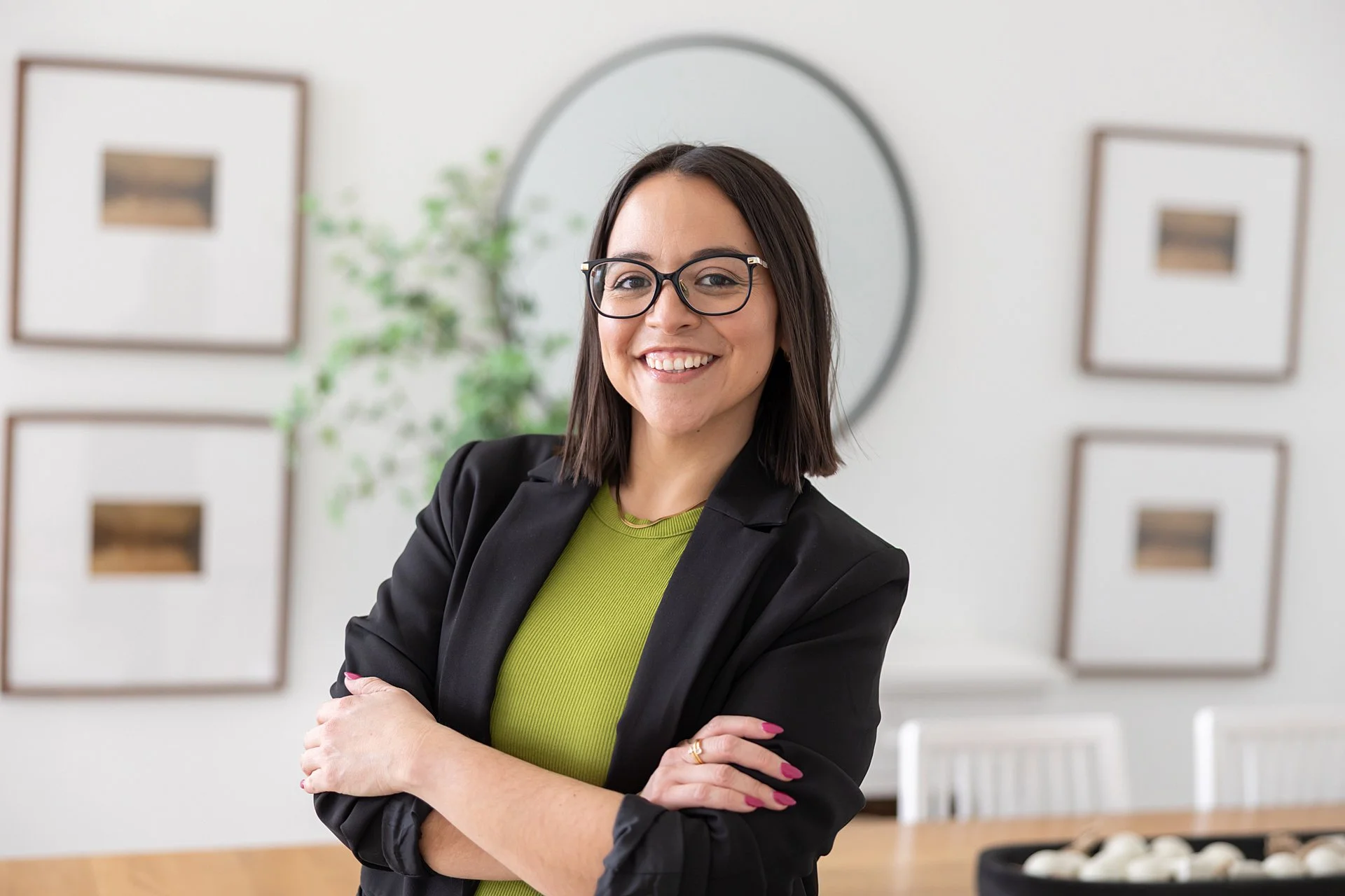 A woman with glasses, shoulder-length dark hair, wearing a black blazer and green top, standing with arms crossed and smiling in a modern, well-lit room with minimalist decor.