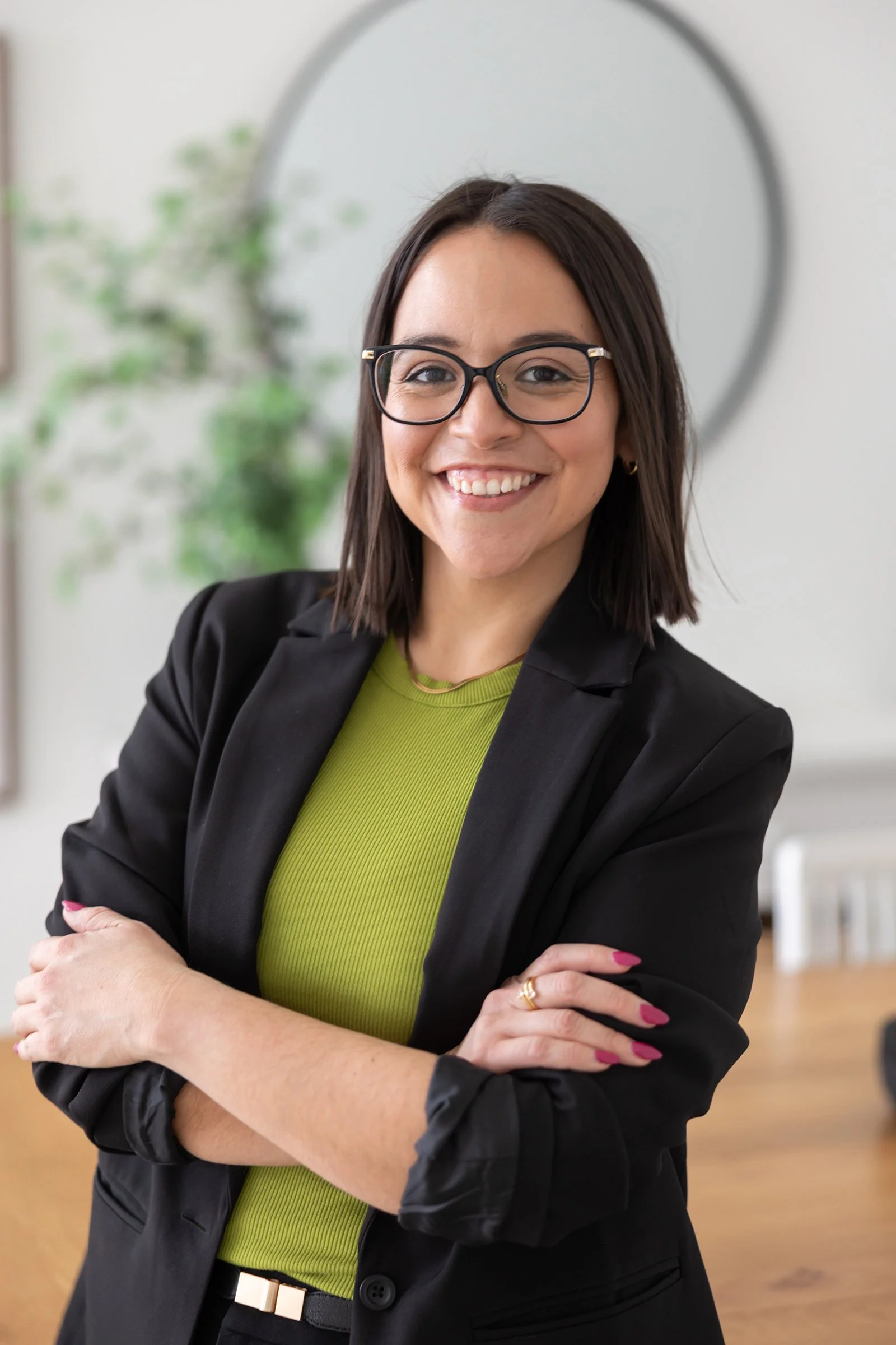 A woman with shoulder-length dark hair, glasses, and a bright smile, wearing a black blazer over a green top, with her arms crossed, standing in a modern room with a round mirror and a plant in the background.