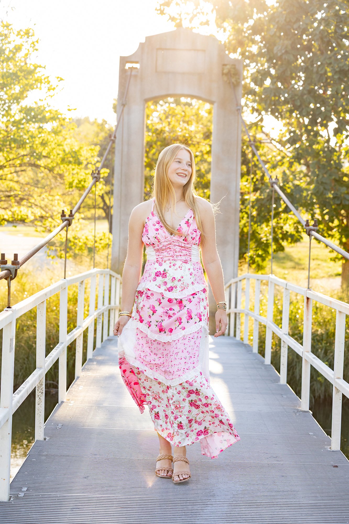 A young woman with long blonde hair walking on a small bridge, smiling and wearing a floral pink and white dress, with trees and sunlight in the background.