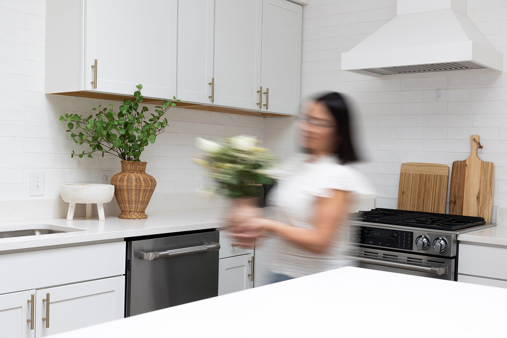 A woman holding a bouquet of flowers in a modern kitchen with white cabinets, a large potted plant, and a stainless steel oven.