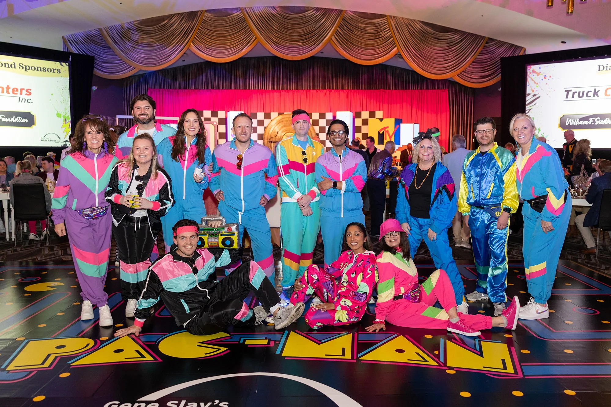 Group of people in colorful 1980s-style athletic outfits posing at an event with a large screen and stage in the background, featuring neon and retro decor.