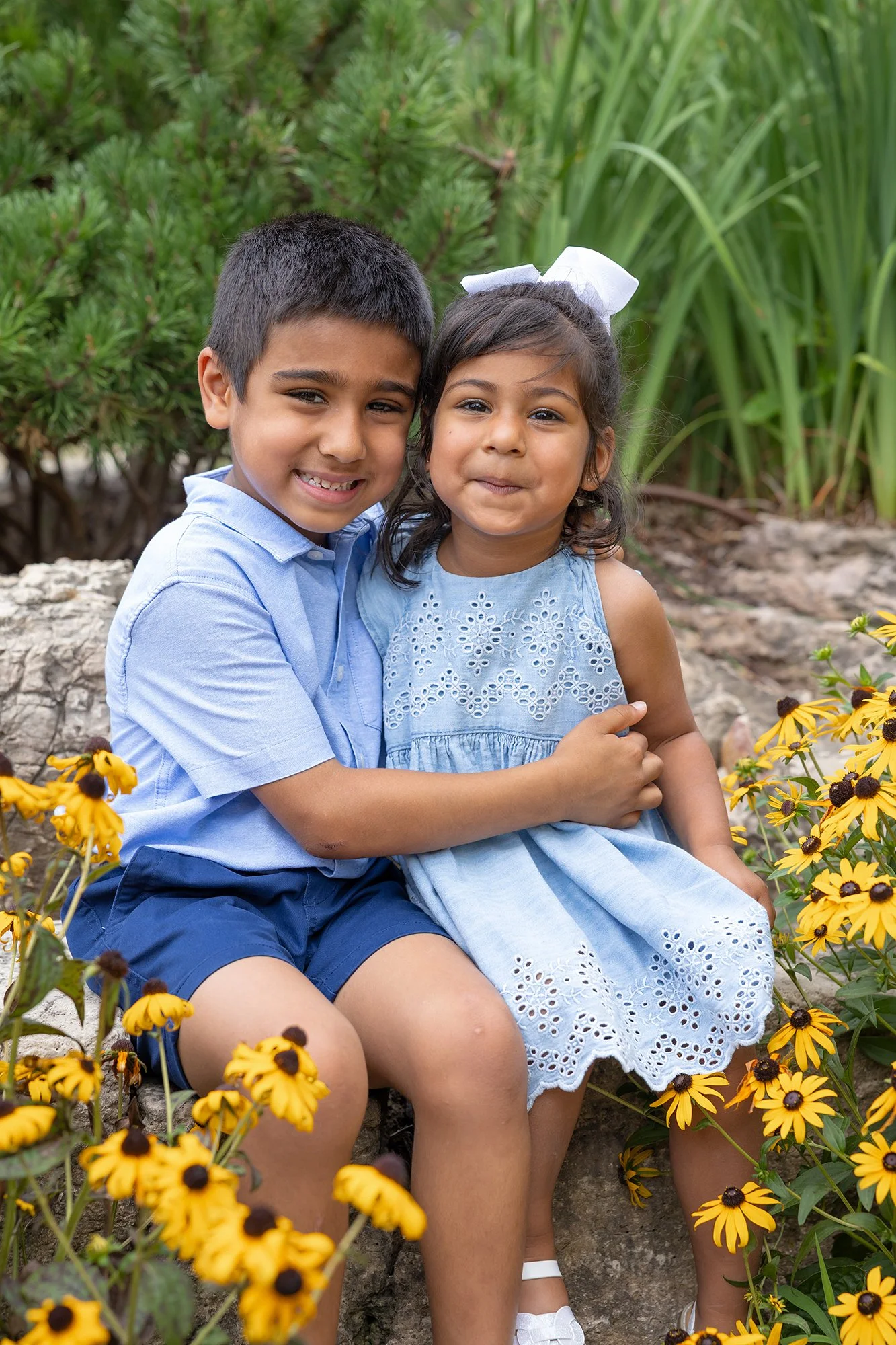 A young boy and girl sitting together on a rock, surrounded by yellow flowers and green foliage, smiling at the camera.