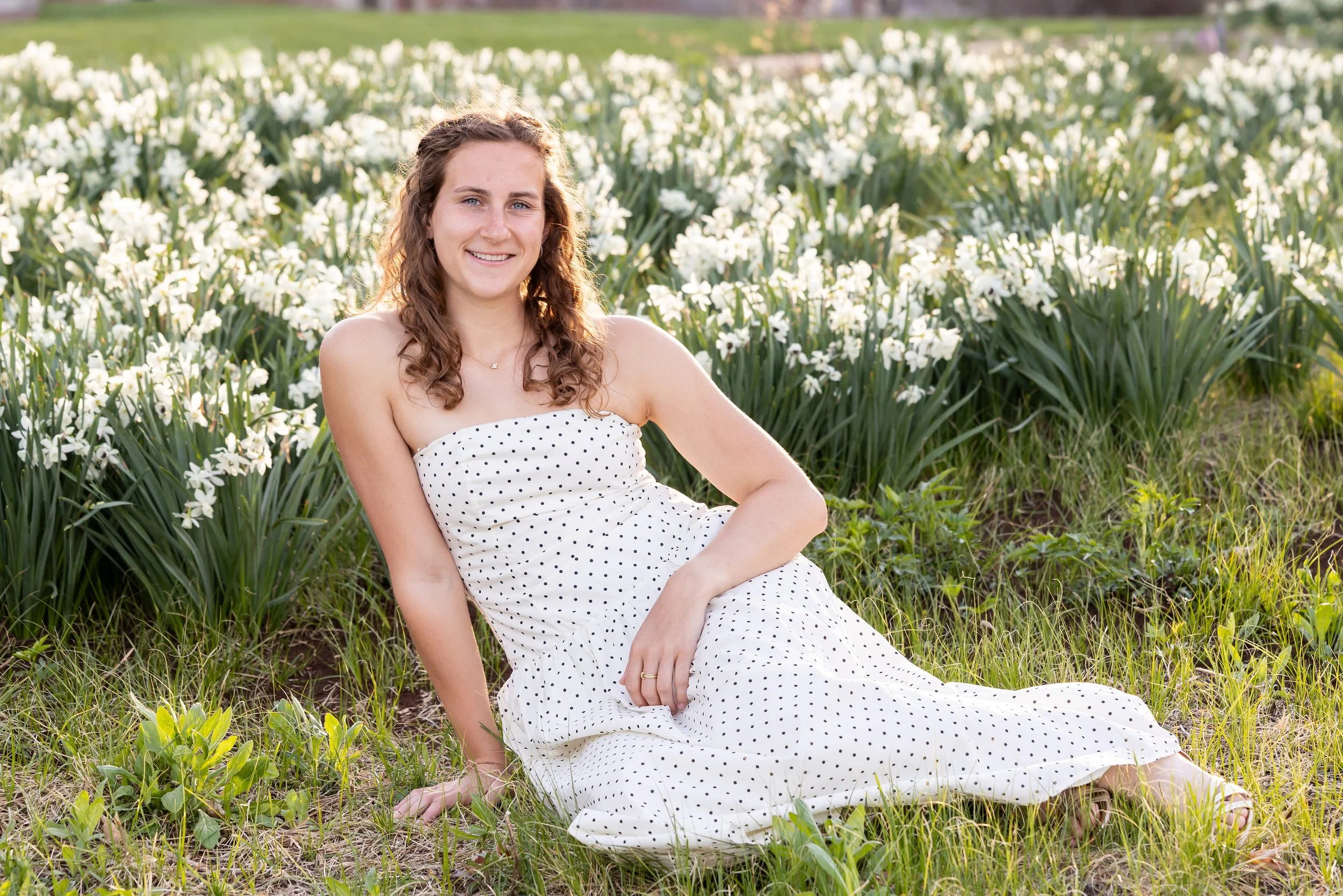 A young woman in a white dress with black polka dots sitting in a field of white flowers and green grass, smiling at the camera.