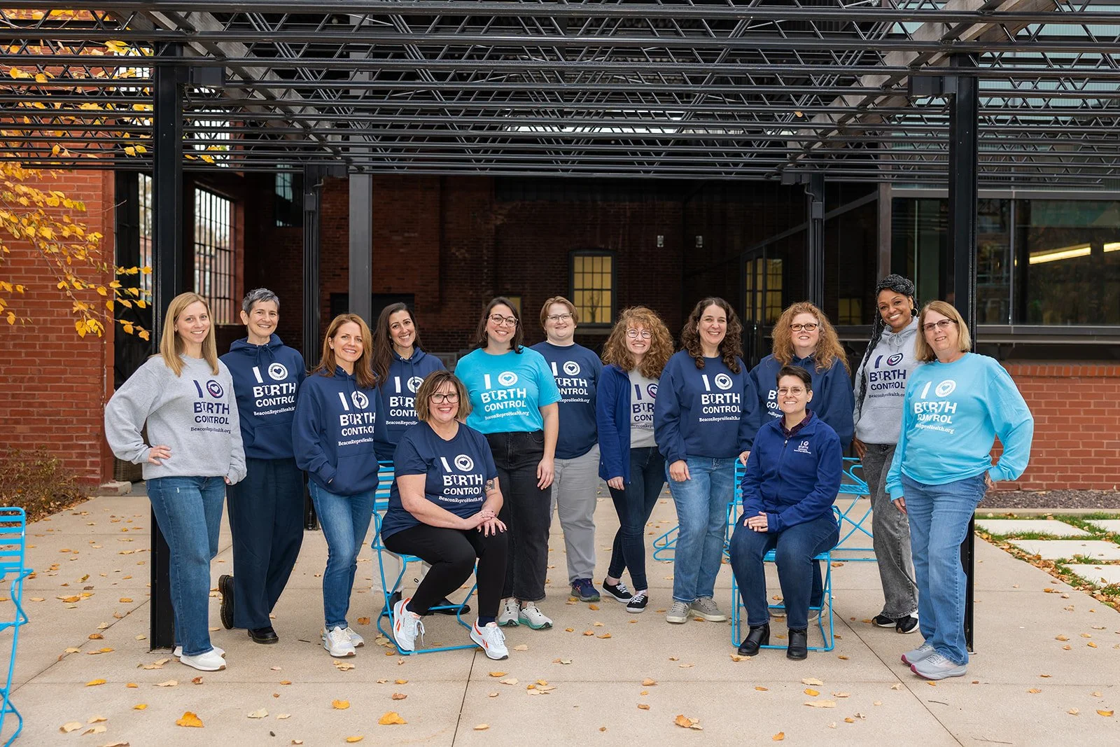 Group of 13 women standing outside, some sitting, all wearing blue or gray hoodies with the text 'I ♥ BIRTH CONTROL' and 'BecomingPermissive.org'
