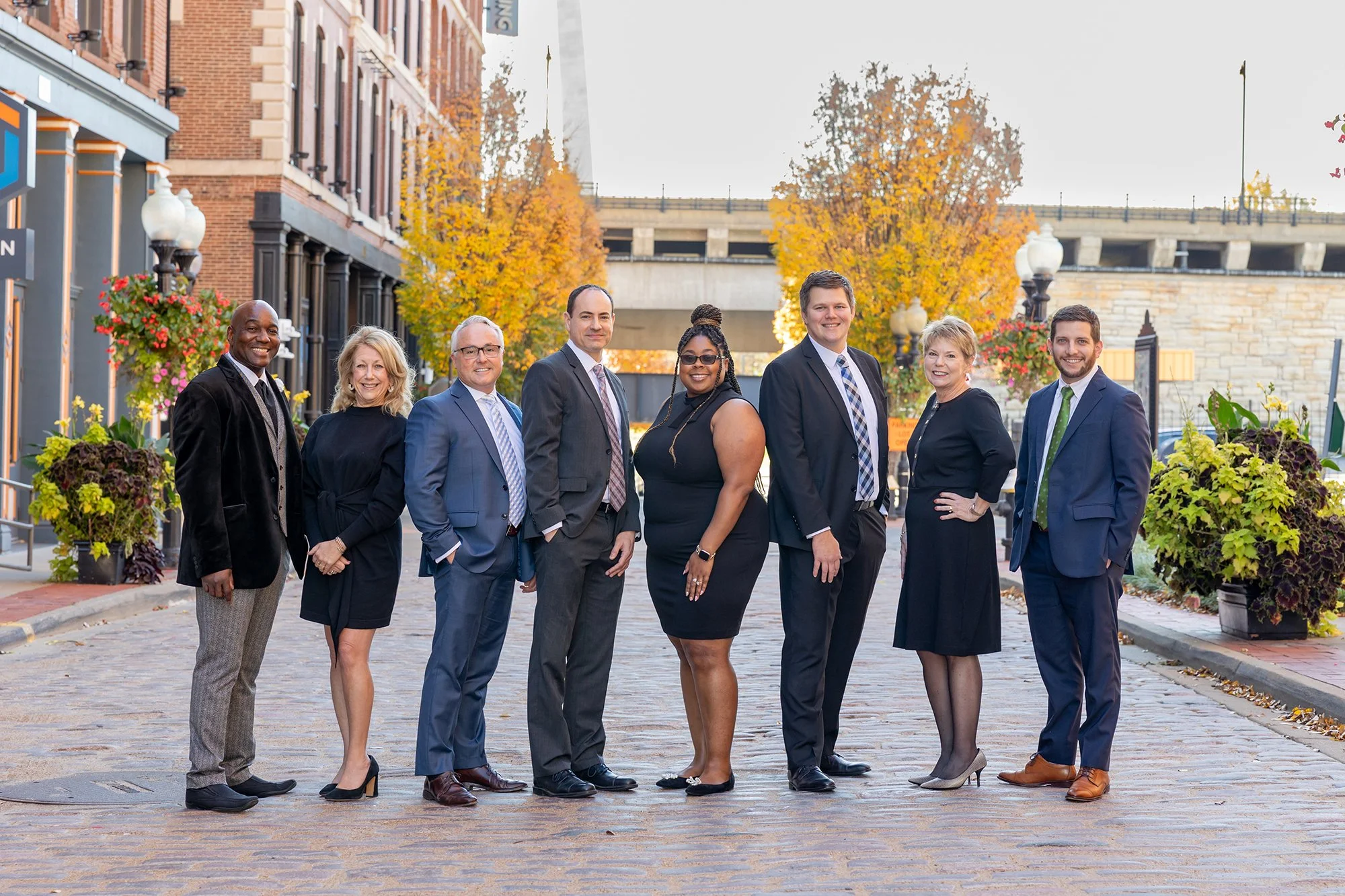 Group of eight diverse professionals standing on a cobblestone street, dressed in business attire, with autumn trees and city buildings in the background.