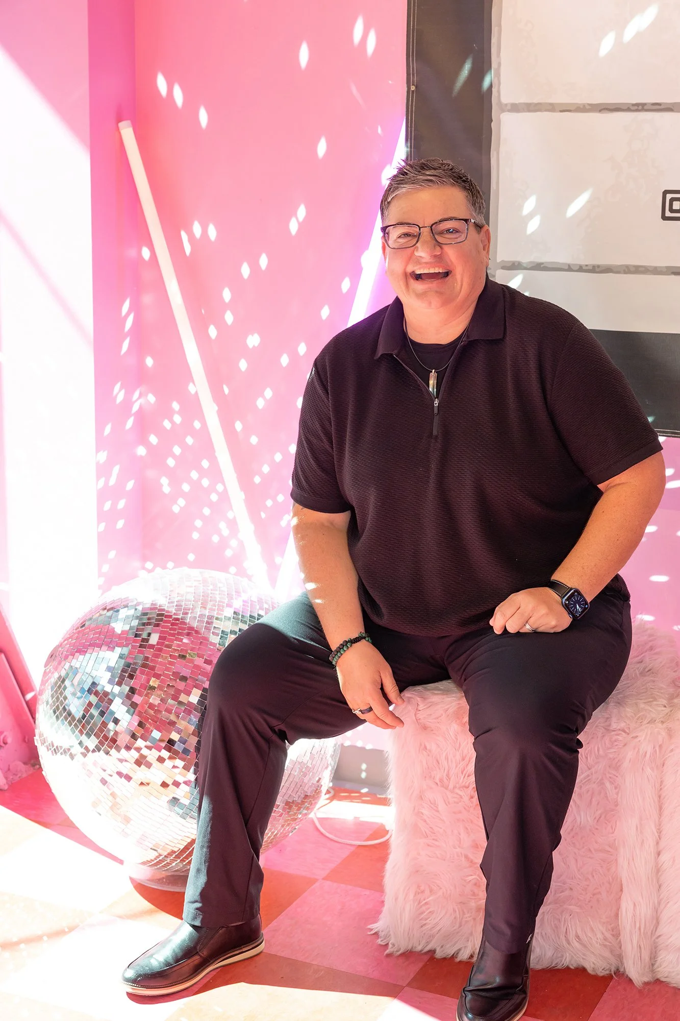 A person with short hair and glasses, dressed in a black polo shirt and black pants, is sitting on a fluffy pink stool. They are smiling, with a disco ball on the floor behind them and pink-colored walls decorated with light patterns.