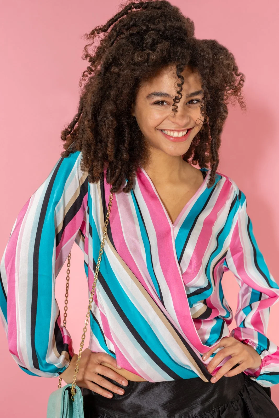 Young woman smiling with curly hair, wearing a colorful striped blouse and black skirt, standing against a pink background.