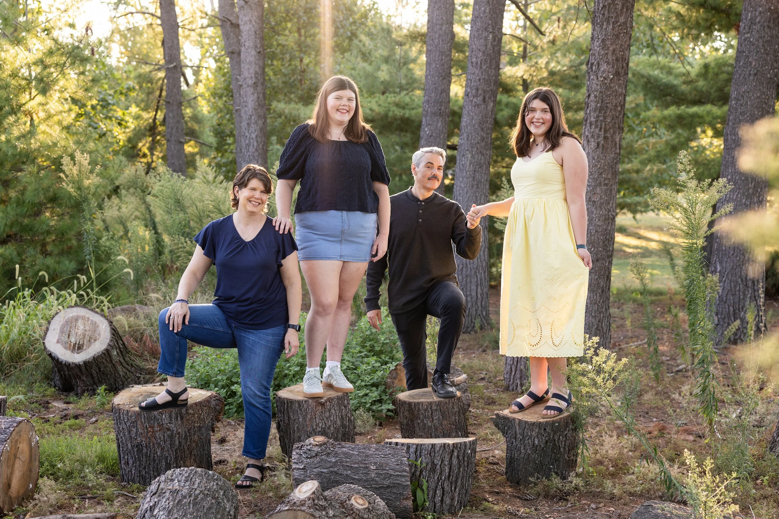 Five people standing and kneeling on tree stumps in a forest, smiling and holding hands. The sunlight filters through the trees, creating a warm ambiance.