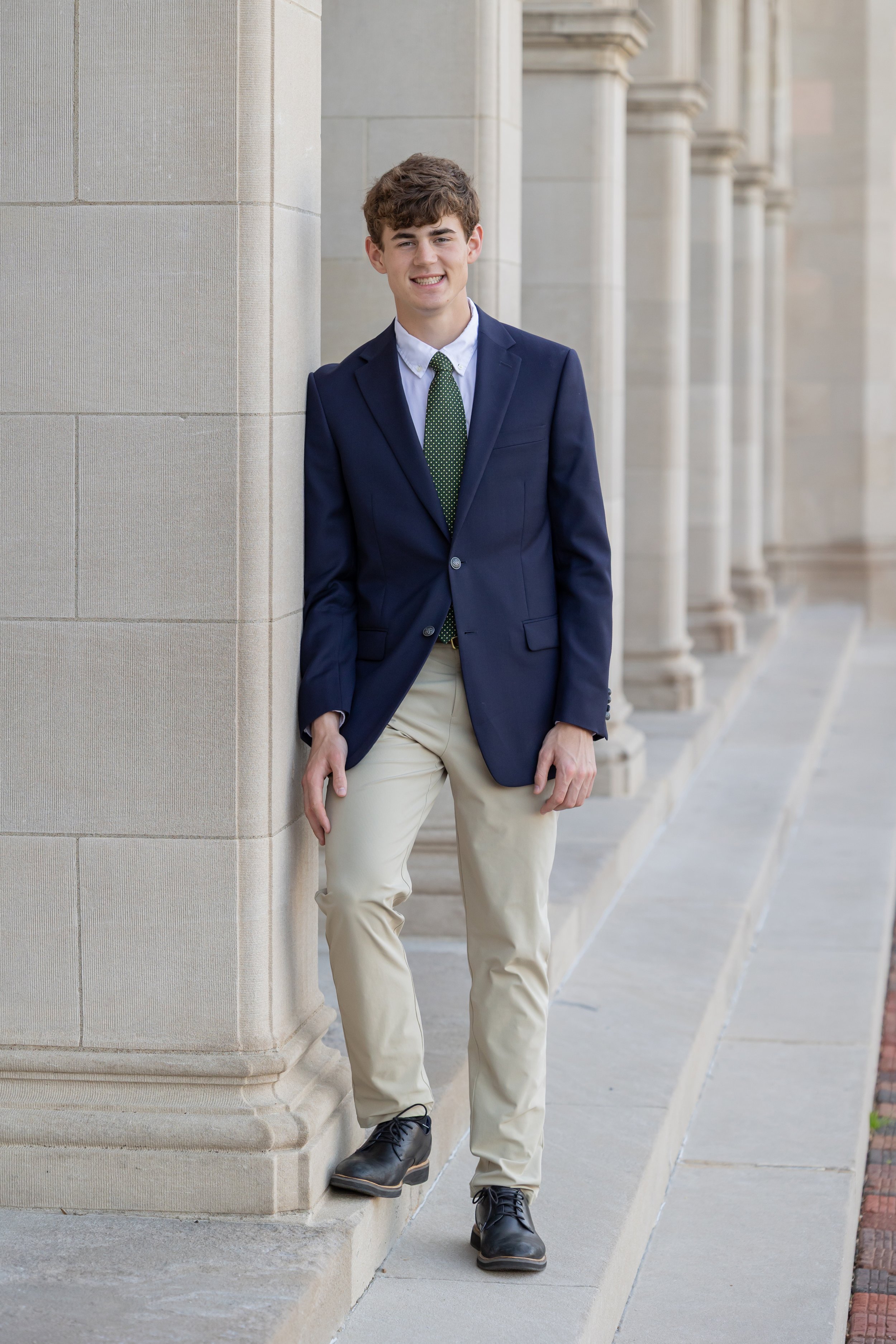 A young man in a navy blazer, white shirt, and green tie leaning against a stone column outside a building