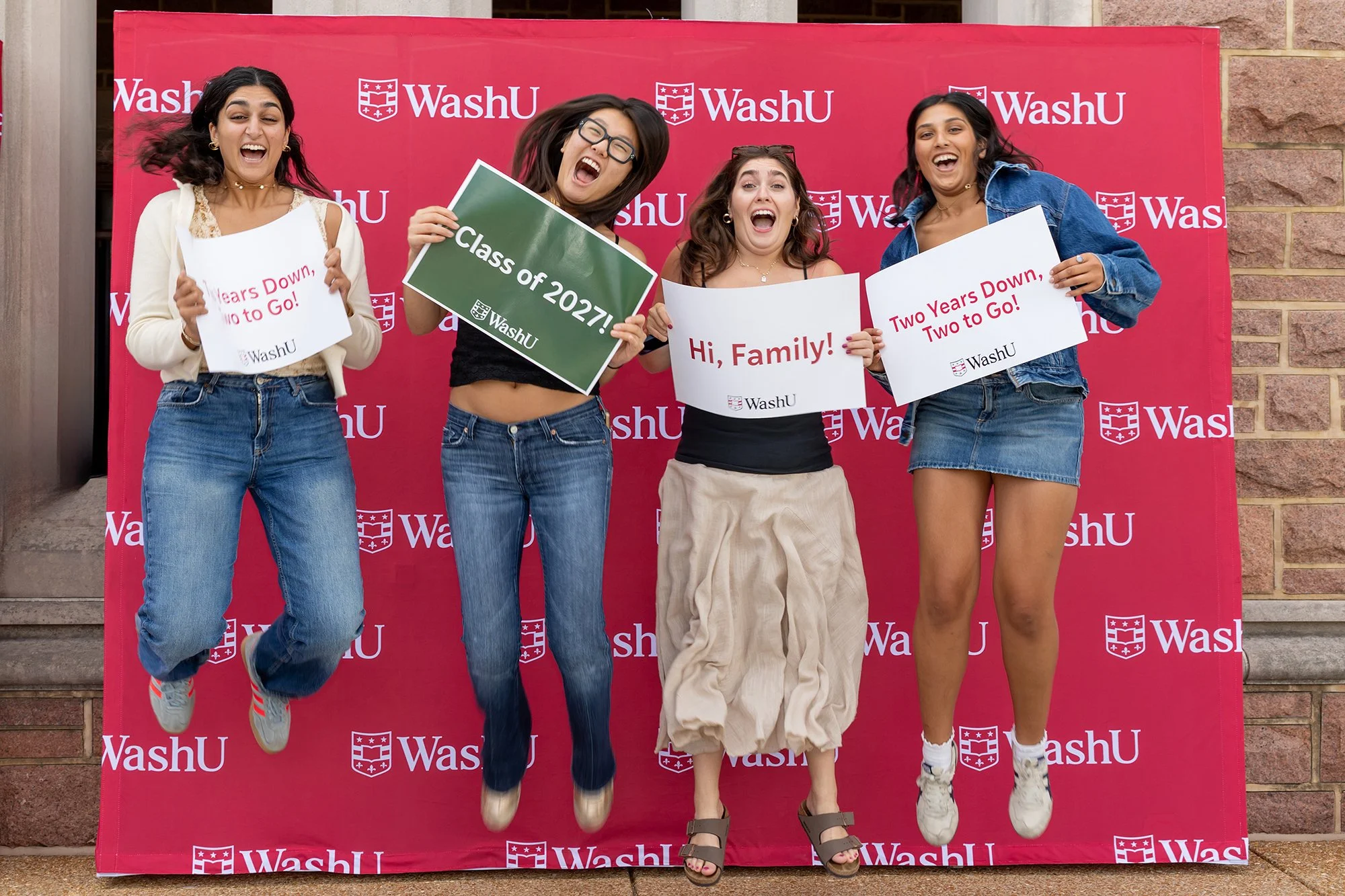 Four young women celebrating graduation at WashU, holding signs that say "Class of 2027!", "Hi, Family!", "Two Years Down, Two to Go!", and one partially visible. They are jumping and smiling in front of a red WashU backdrop.