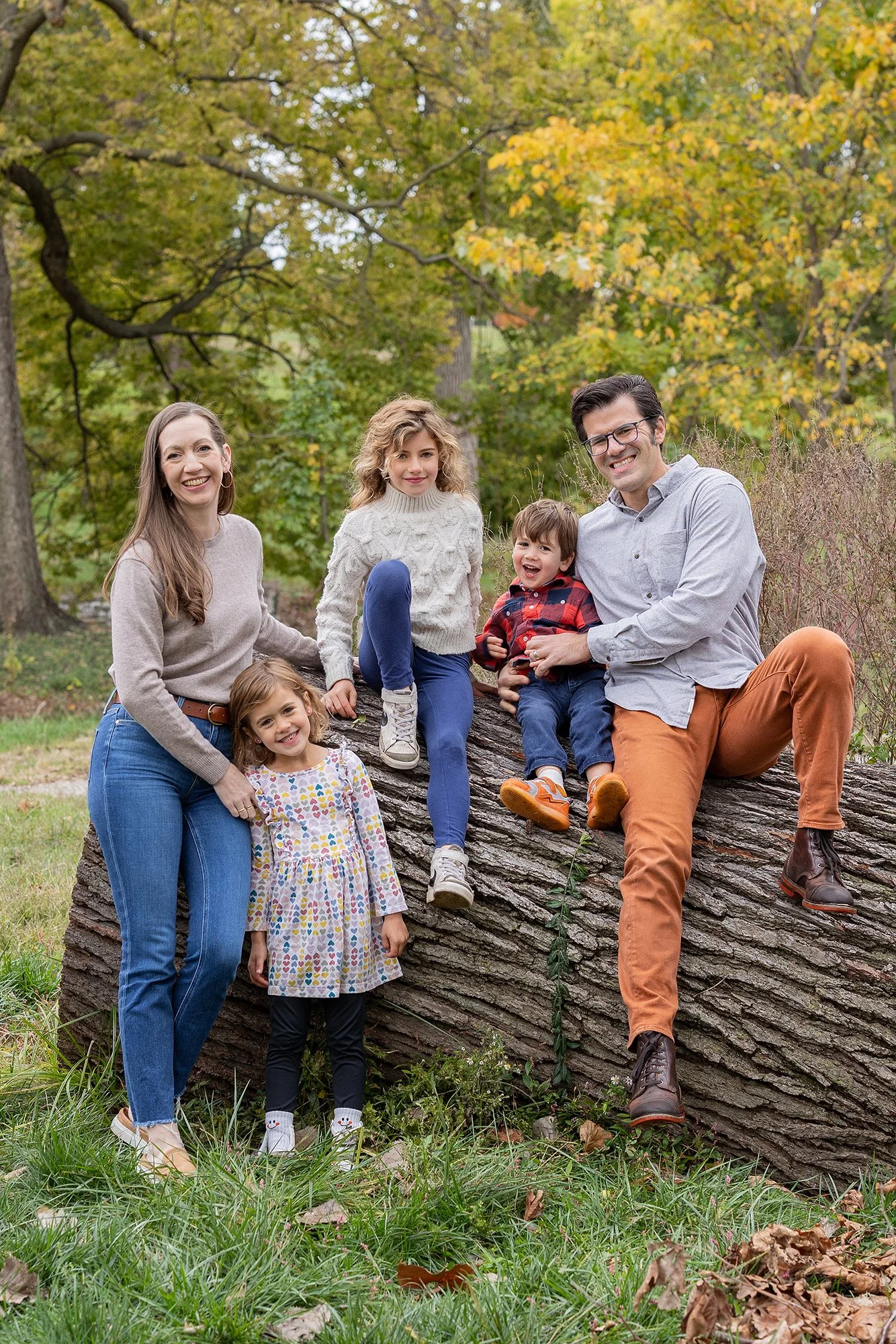 Family of six enjoying outdoor fall scenery, sitting on fallen tree in a park with autumn-colored leaves.