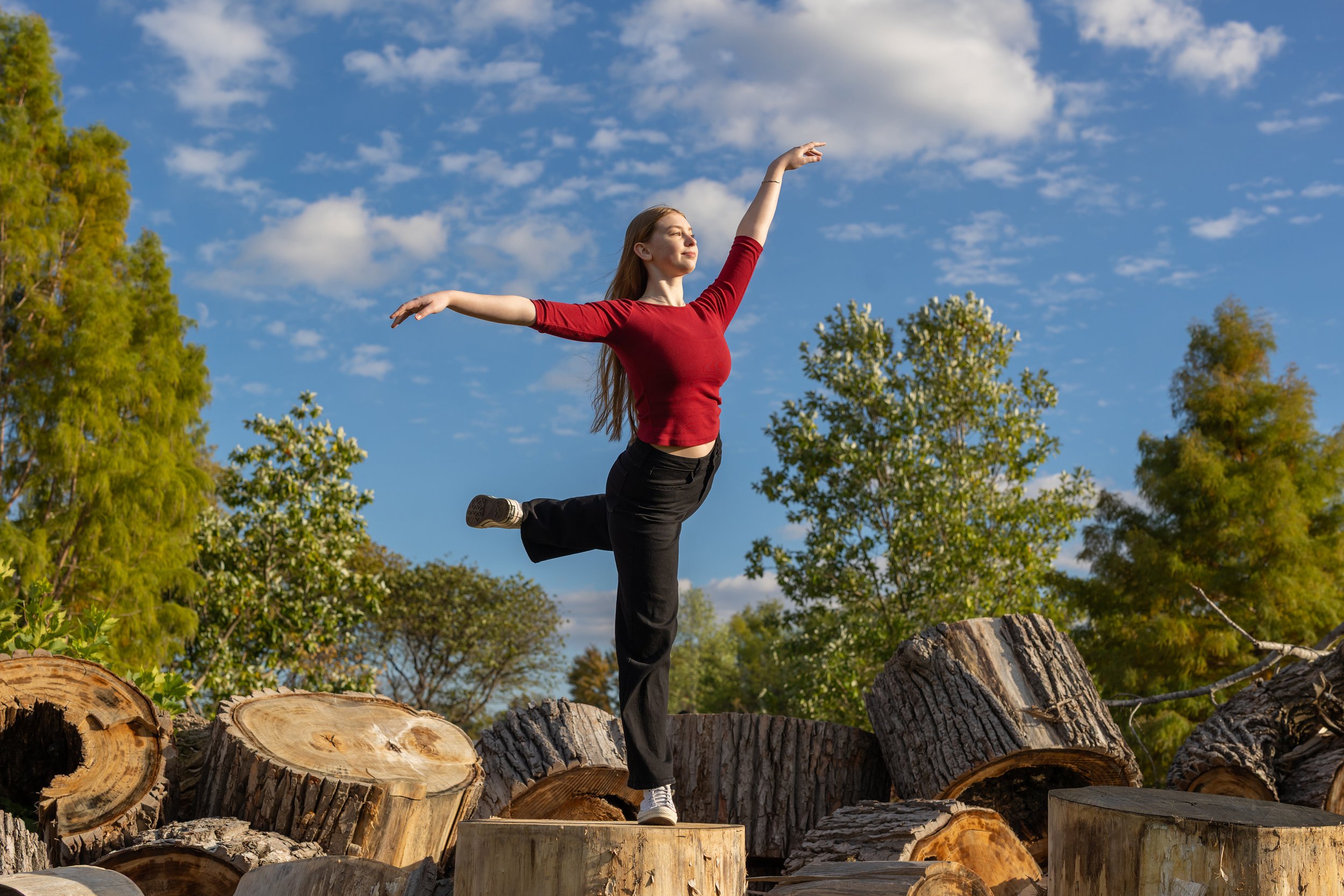 A woman with long hair in a red top and black pants balancing on one leg on a tree stump outdoors, with trees and a partly cloudy sky in the background.
