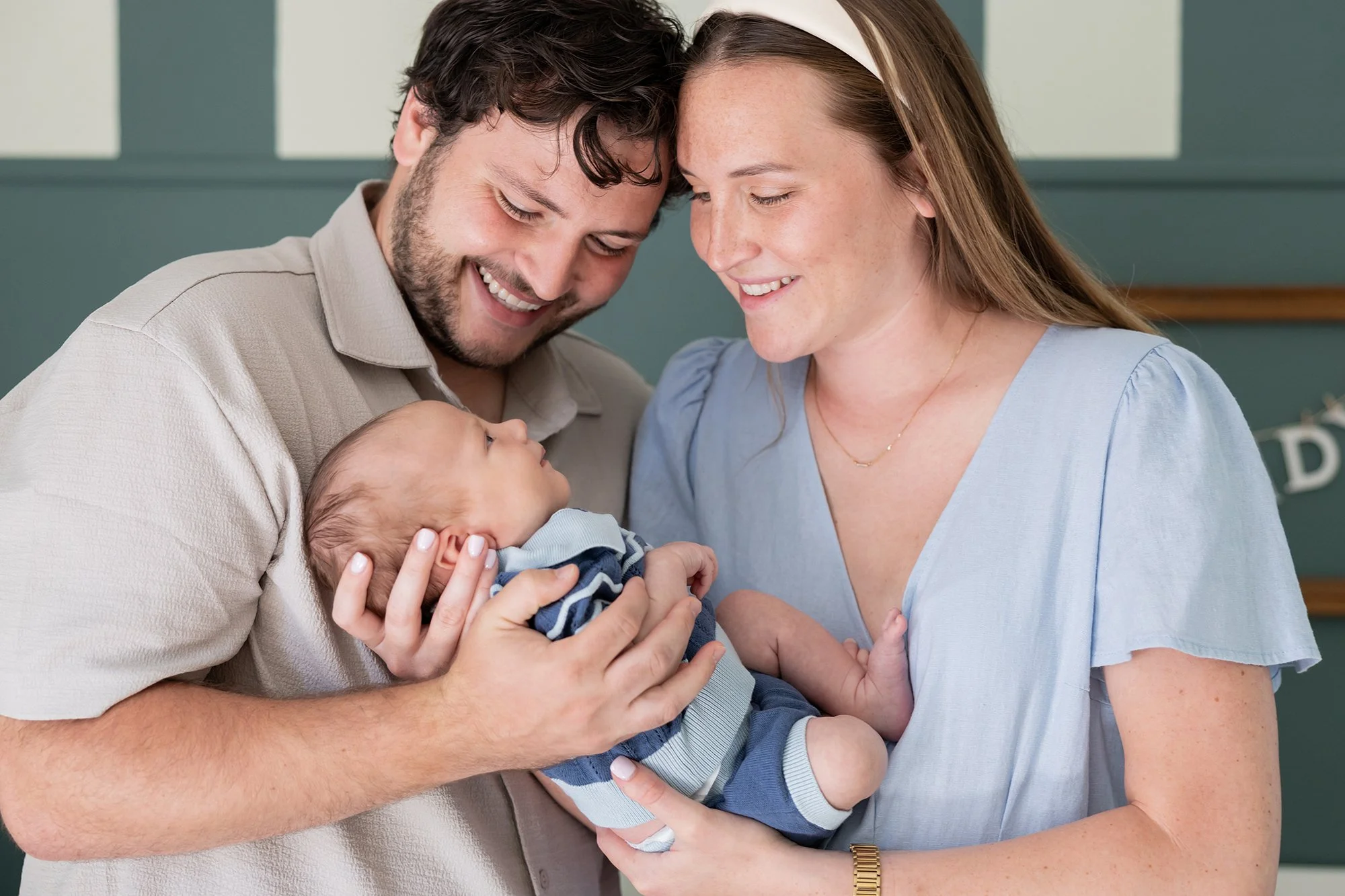 A smiling man and woman are holding a newborn baby, looking at each other and the baby affectionately.