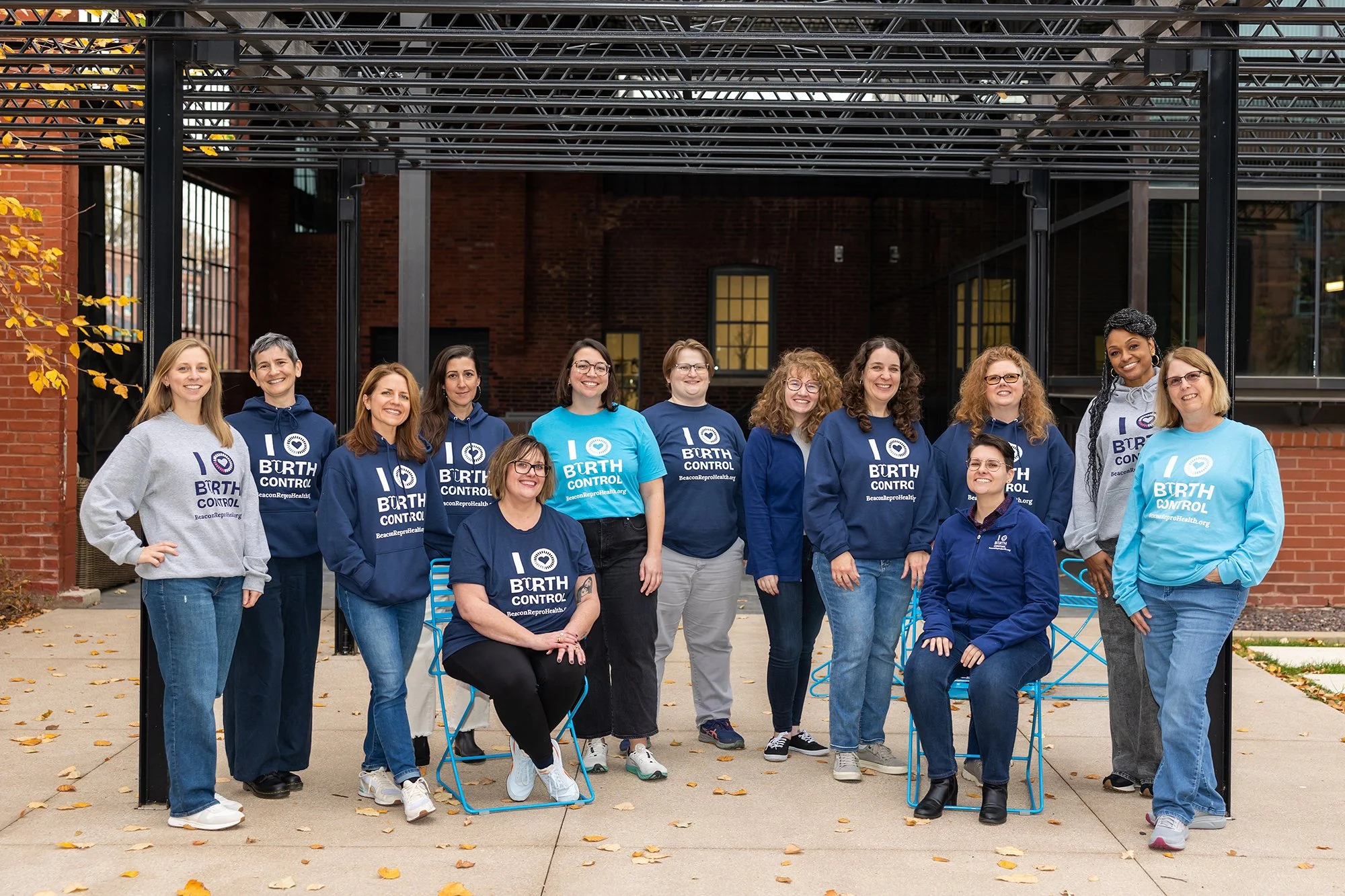 Group of women wearing matching shirts and hoodies that say "I ♥ Birth Control" standing and sitting outdoors on a sidewalk with autumn leaves, brick buildings, and metal structures in the background.