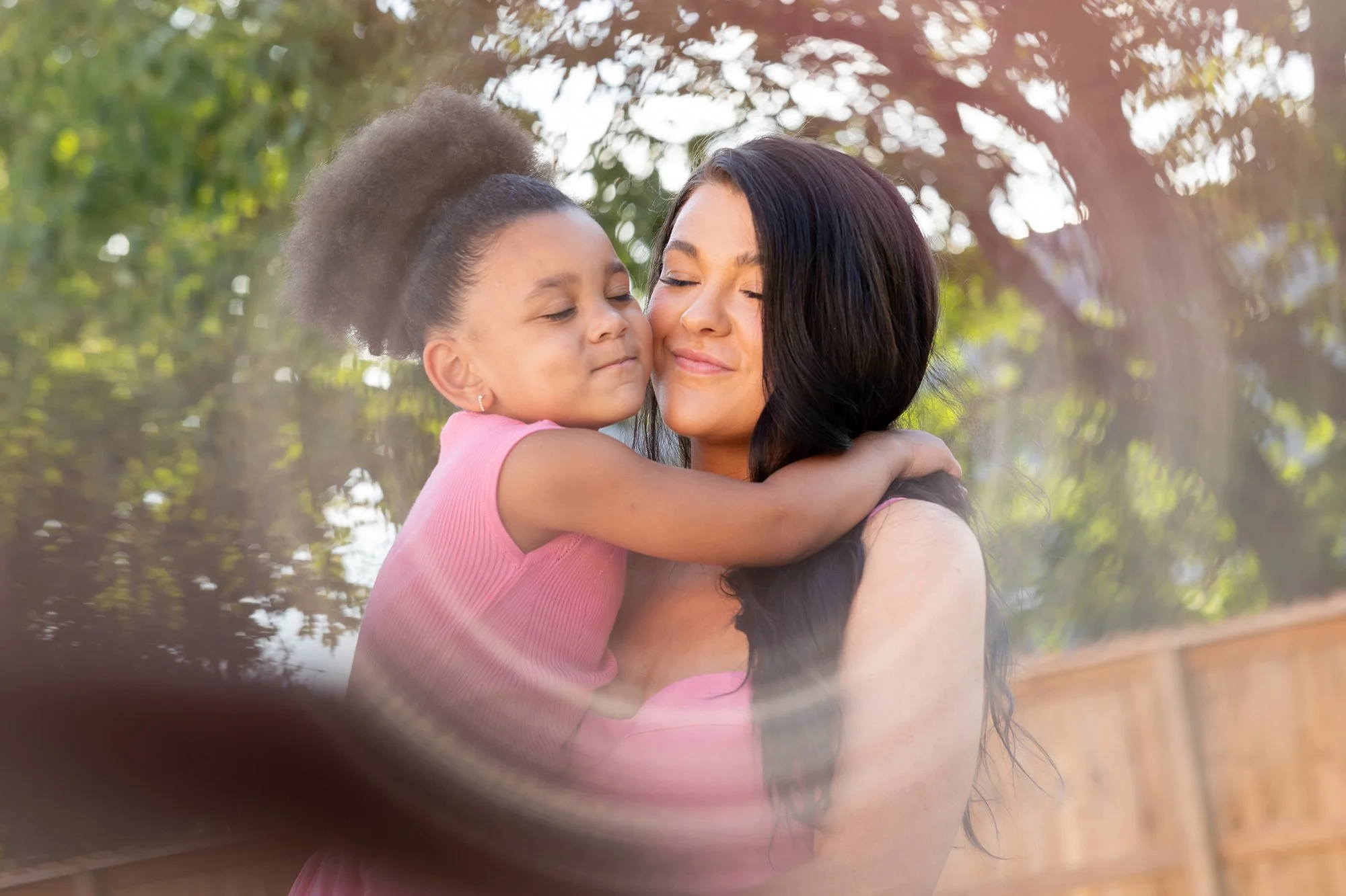 A young girl with curly hair and a woman with long dark hair share a loving hug outdoors on a sunny day, with trees and a wooden fence in the background.