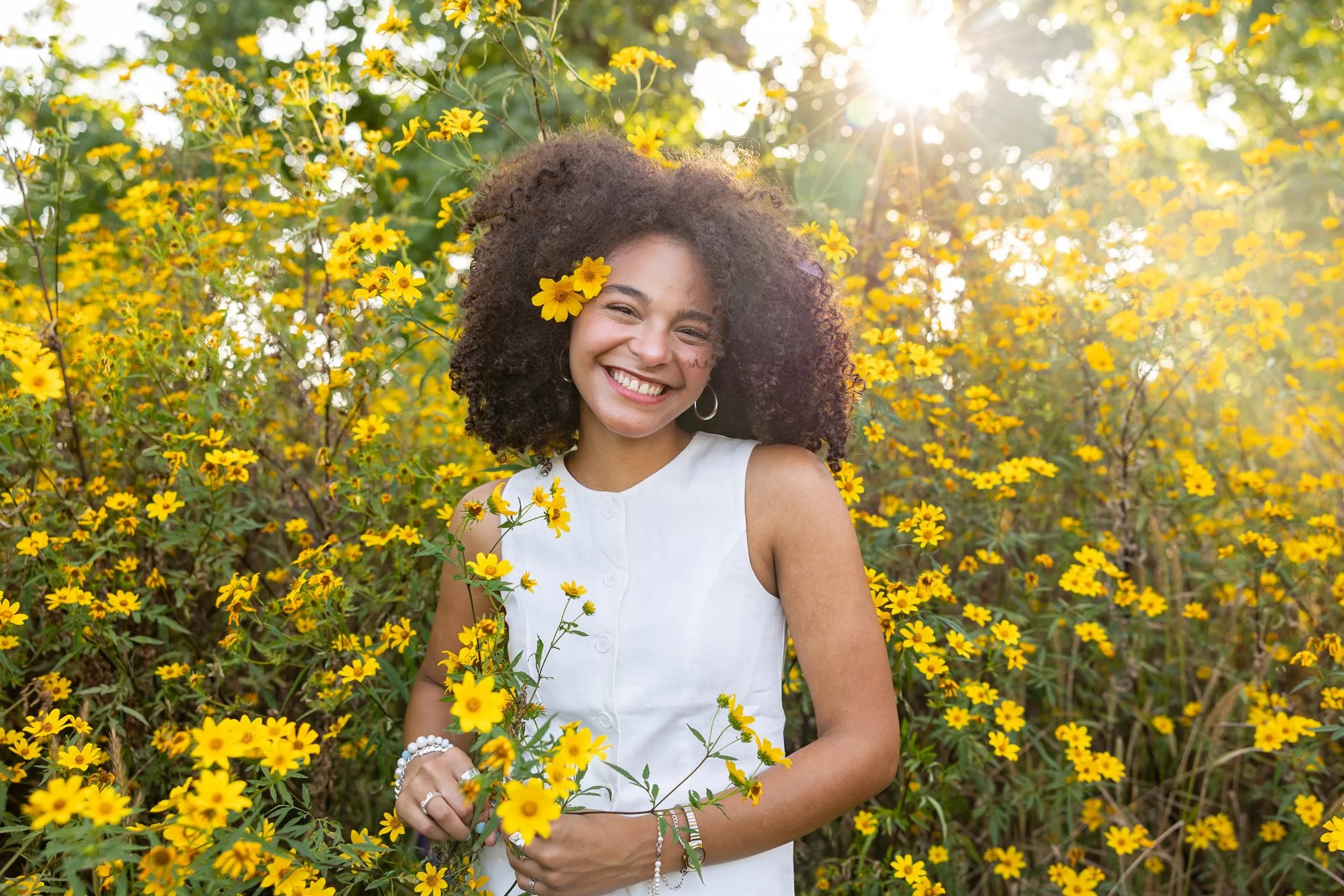A young woman with curly hair and a yellow flower in her hair smiling among yellow flowers in a sunny outdoor setting.