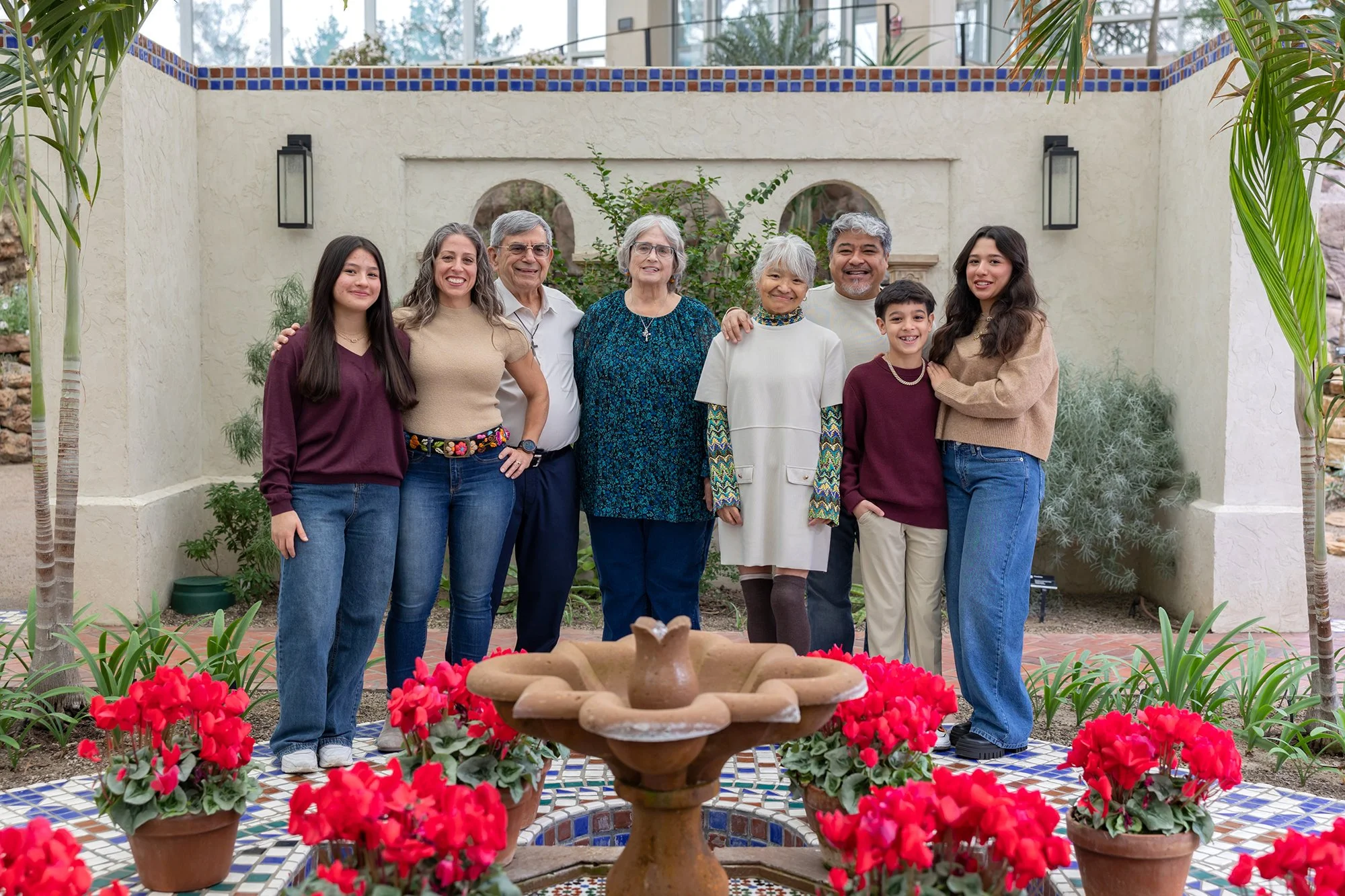 A multigenerational family of nine people standing together in a garden courtyard, smiling for the photo, with a decorative fountain and vibrant red flowers in the foreground, and a Mediterranean-style wall with arches and greenery in the background.