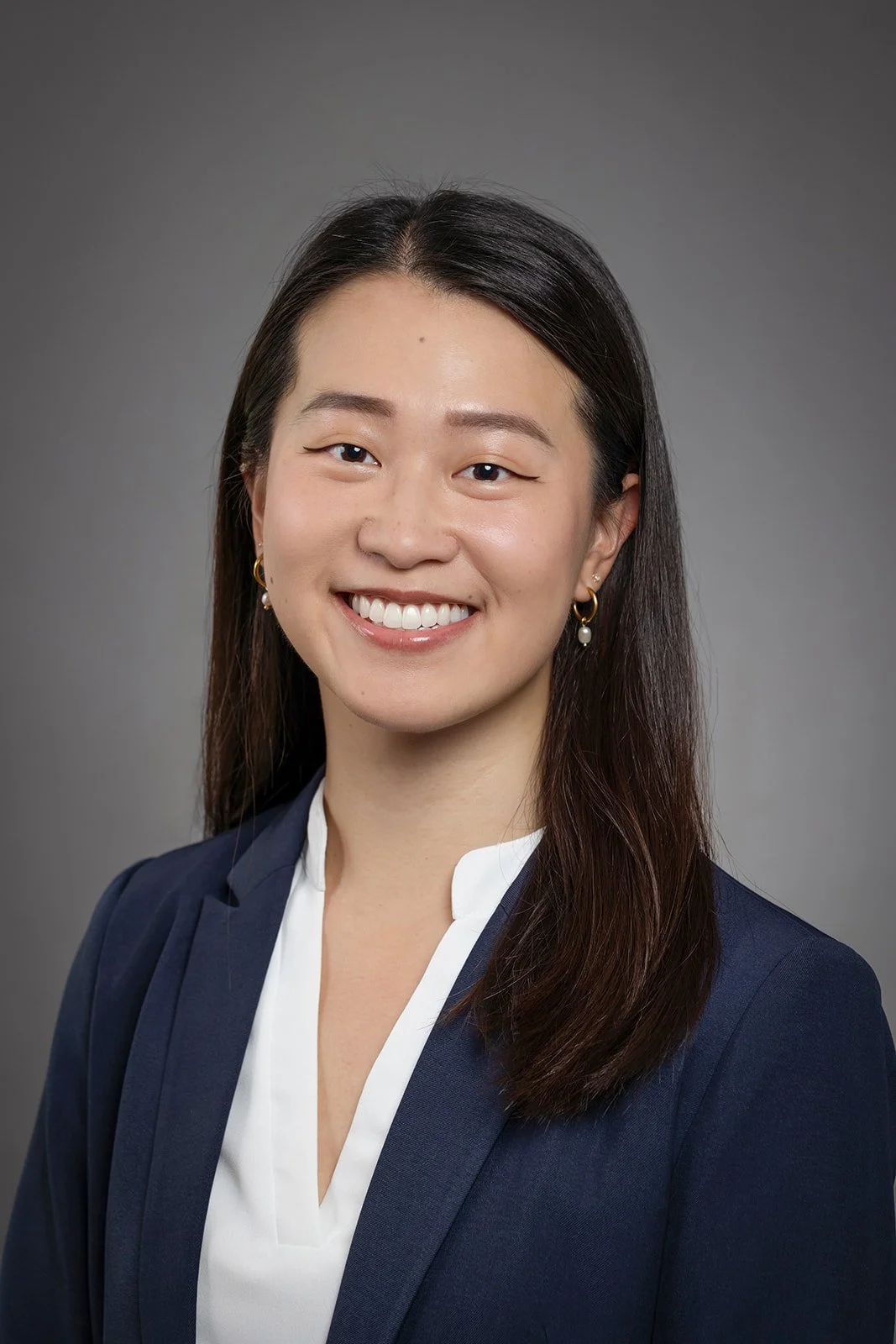 Professional headshot of a woman with long dark hair, wearing a navy blazer, white blouse, gold hoop earrings with pearl details, smiling against a gray background.