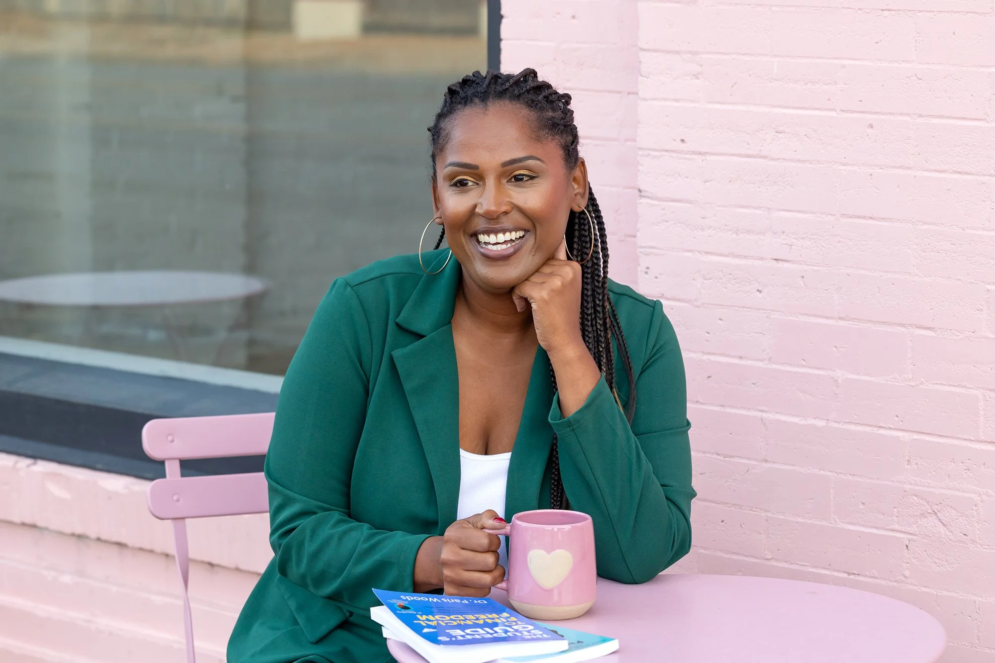 A woman with braided hair, wearing a green blazer, sitting at a pink outdoor table with a pink mug and a book, smiling and resting her chin on her hand, against a pink brick wall.