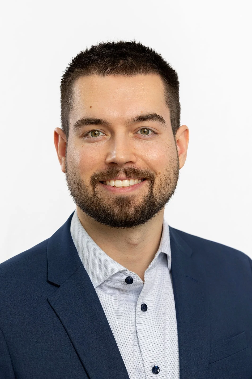 A young man with short dark hair, a beard, and green eyes smiling, wearing a dark blue suit jacket and a light-colored button-up shirt, against a plain white background.