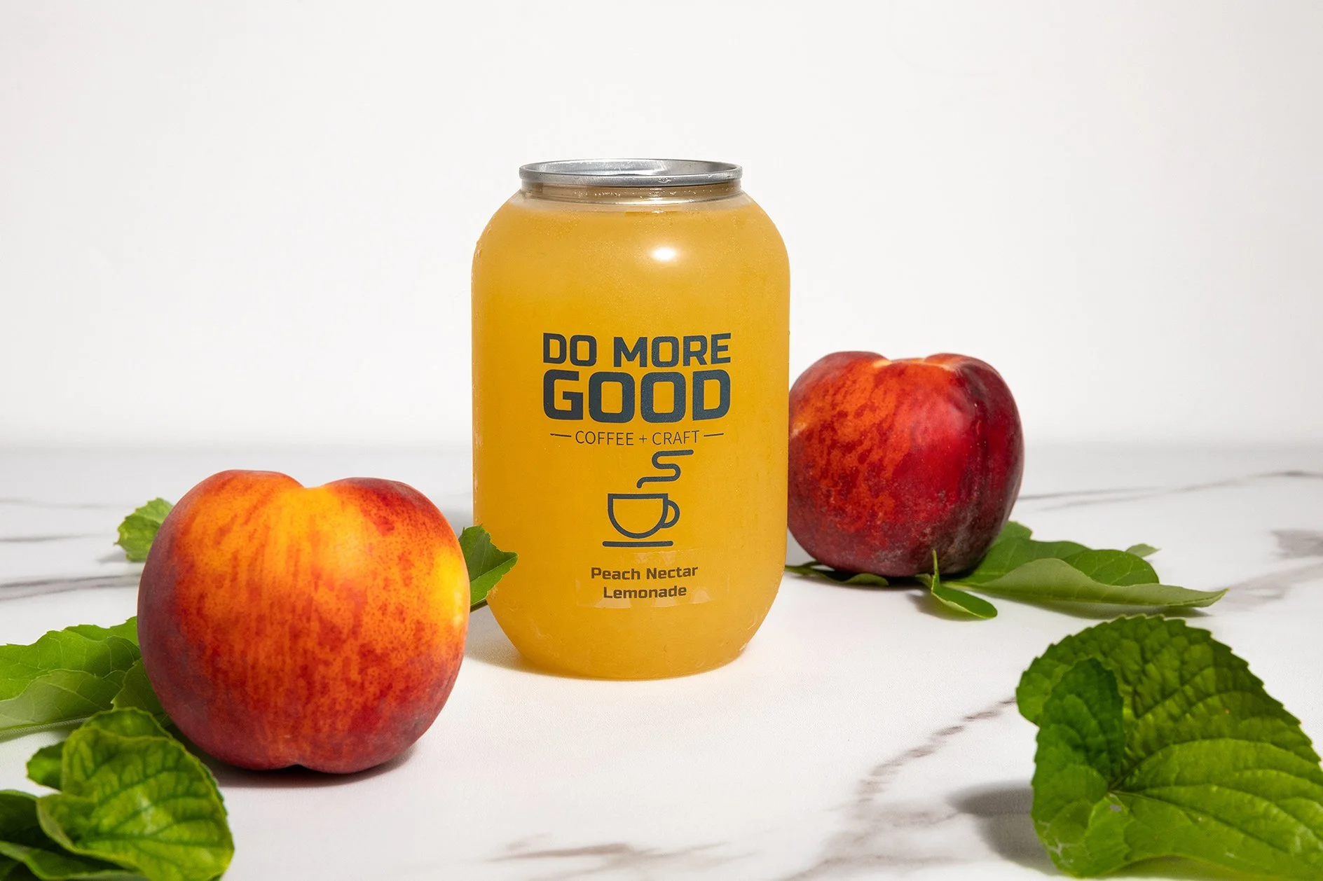 Can of peach nectar lemonade labeled "Do More Good Coffee + Craft" surrounded by peaches and green leaves on white background.