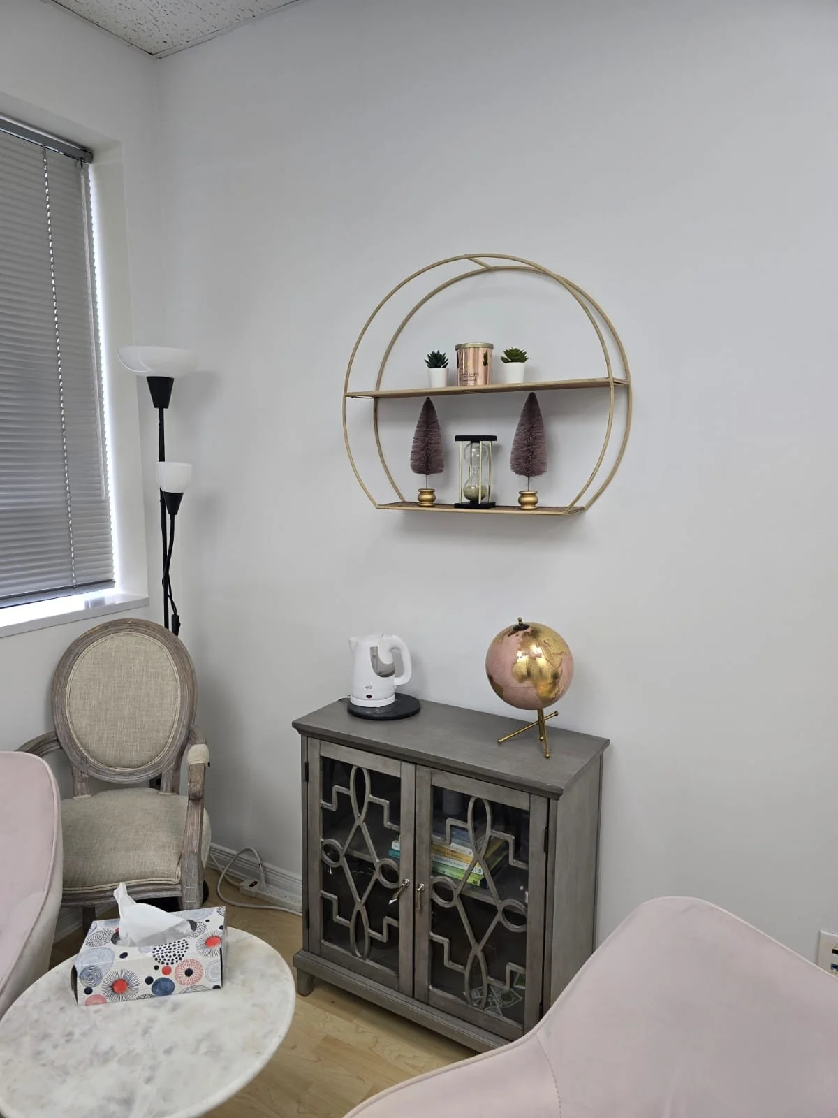 A corner of a room with a window, a tall floor lamp, a beige armchair, a small marble table with a decorative tissue box, a gray cabinet with a globe and electric kettle on top, a wall-mounted round shelf with plants and decorative items, and a white wall.