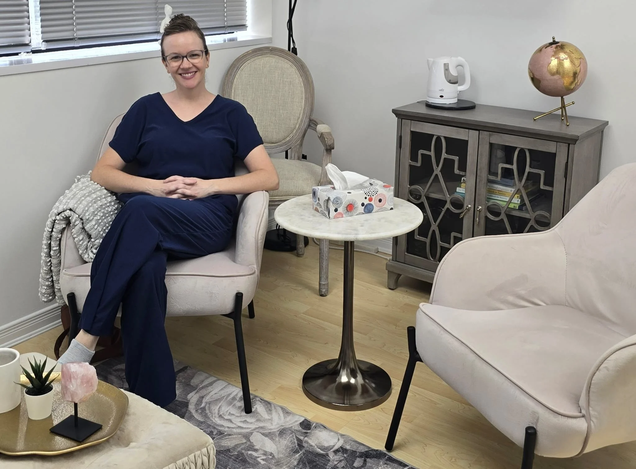A woman sitting on a beige armchair in a room with white walls, smiling at the camera, wearing glasses and navy clothing. There is a small round table with decorative items including succulents and a pink crystal in front of her, and a white tissue box on a small marble-topped side table nearby. A gray painted cabinet with books and a white electric kettle is against the wall, with a decorative globe on top.