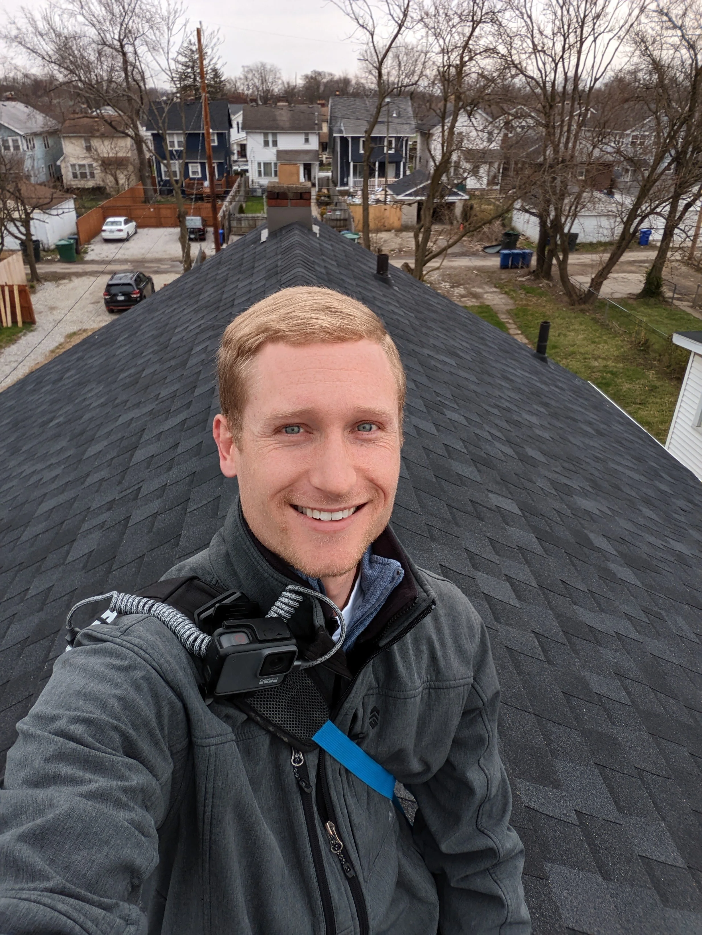A man takes a selfie on the roof of a house with a smartphone camera, smiling, with a neighborhood of houses and trees in the background.
