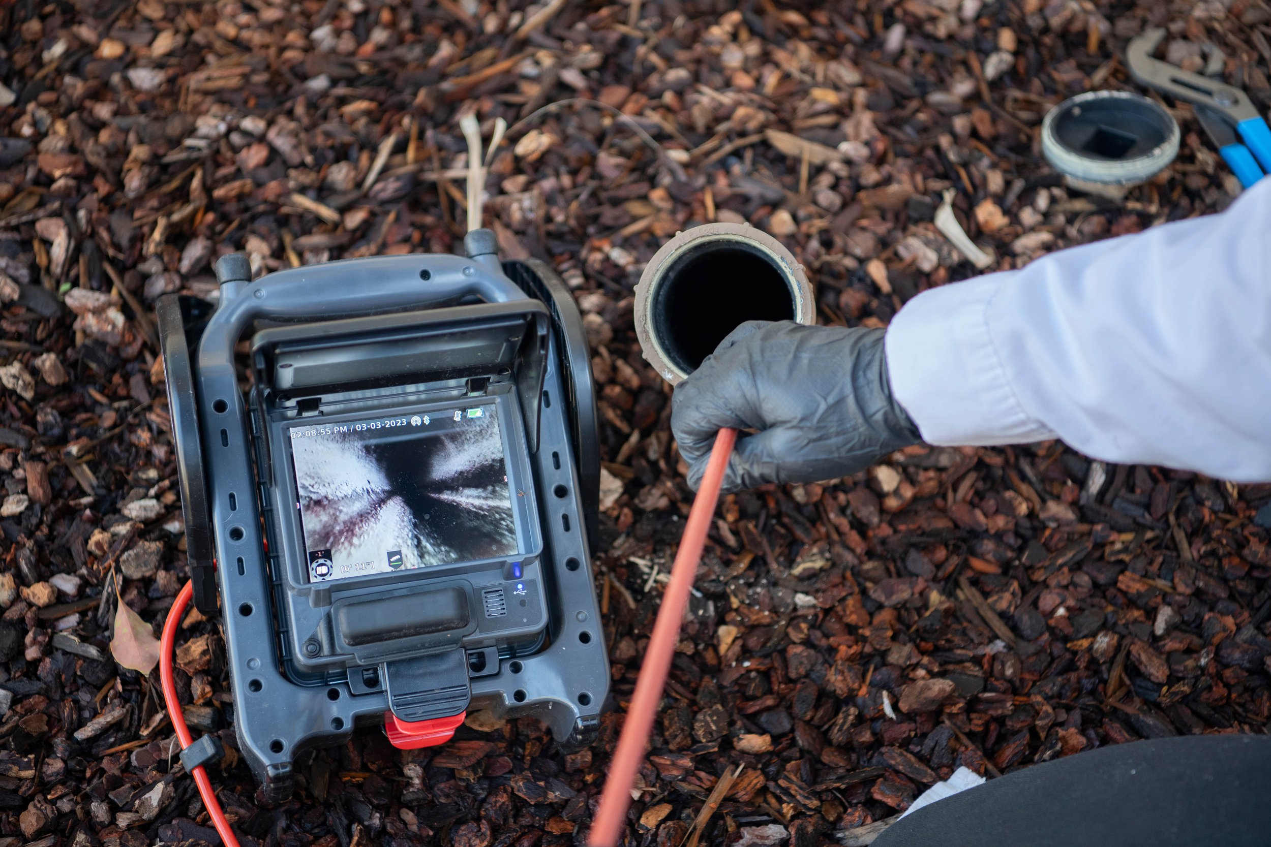 A person in a white coat and black gloves using a pipe inspection camera to examine a pipe on a ground covered in brown bark mulch.