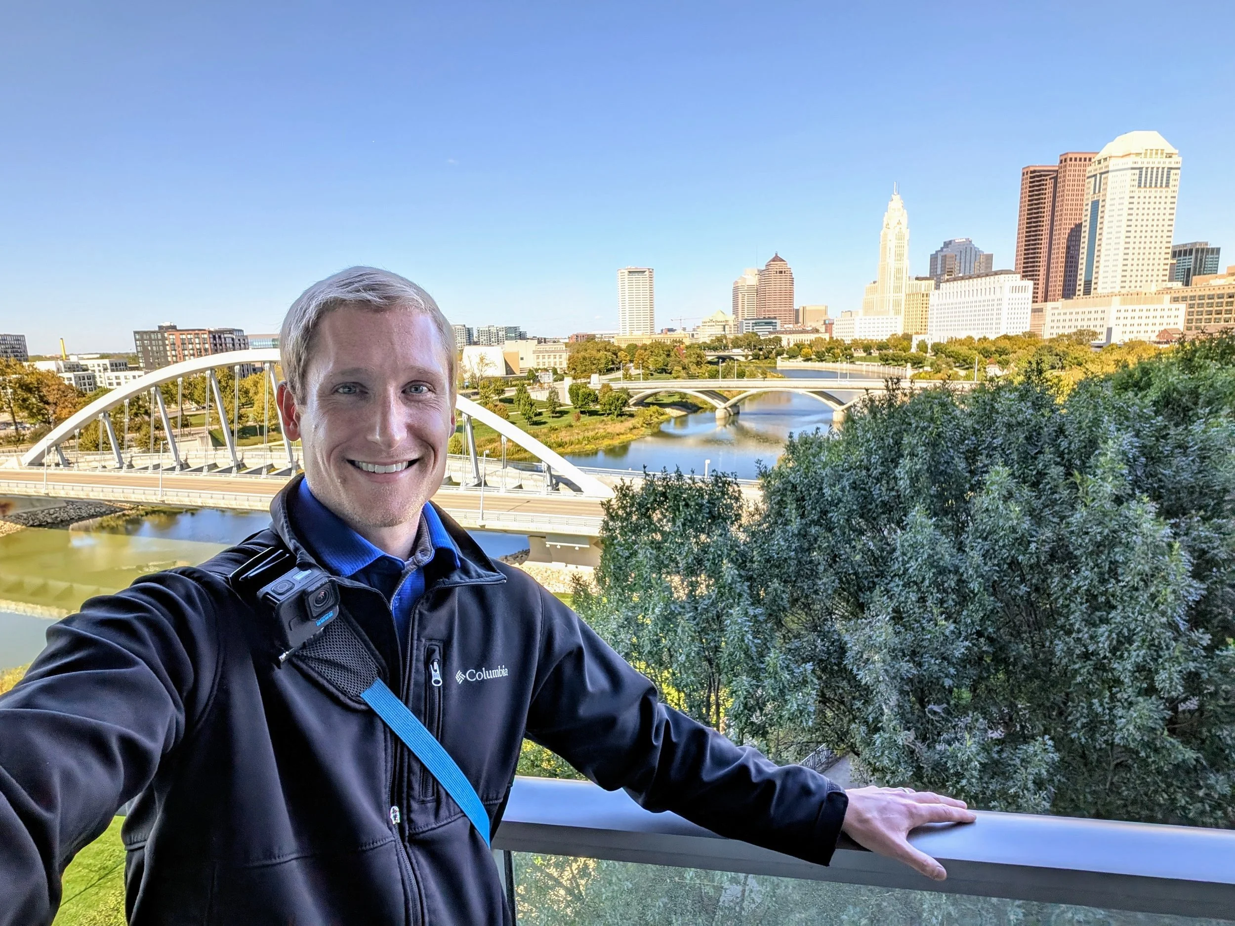 A man taking a selfie on a balcony with a city skyline, including a river and a bridge, in the background on a clear day.