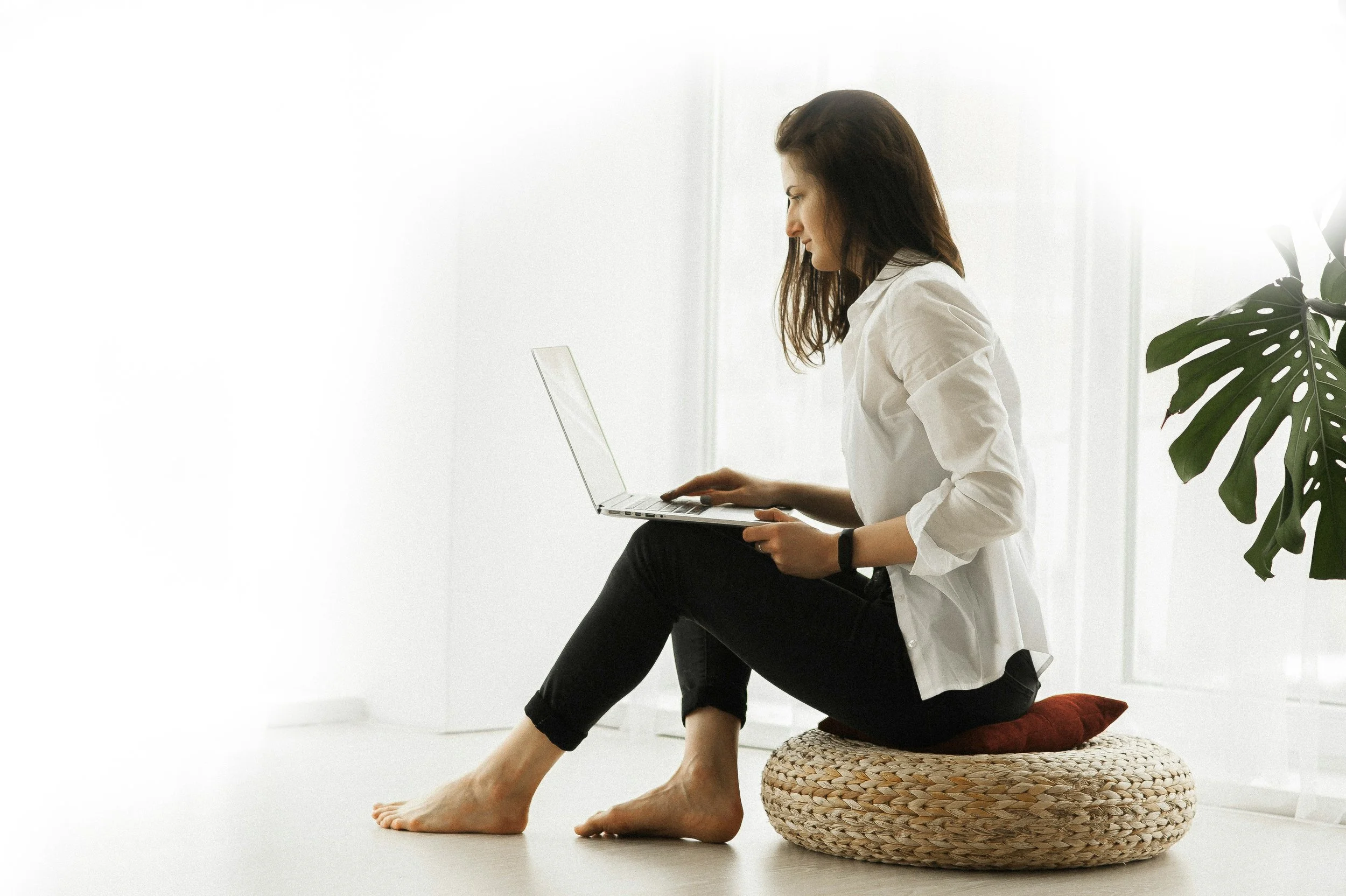 Woman sitting on a woven cushion using a laptop in a bright, minimalist room with a large plant nearby.