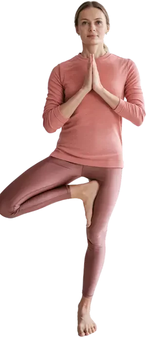 A woman in a pink long-sleeve shirt and matching leggings is practicing yoga in a tree pose, standing on one leg with her hands together in a prayer position at her chest.