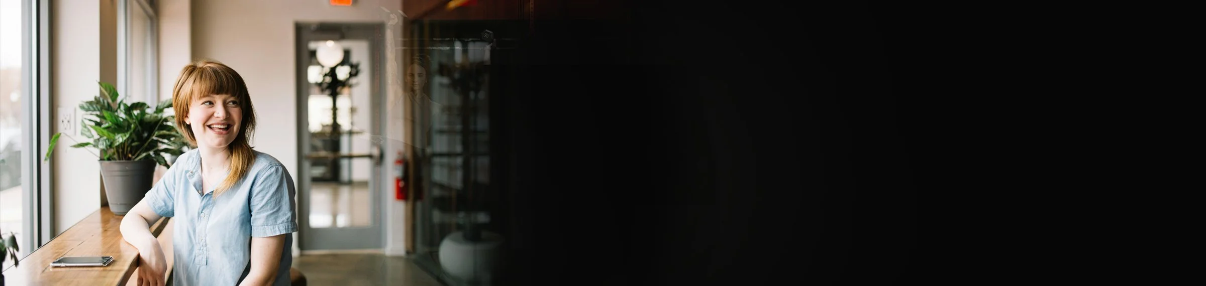 A woman with long brown hair and a light blue shirt sitting at a wooden counter near a window with potted plants, smiling.