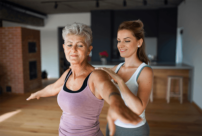 A young woman assisting an elderly woman with a stretch in a modern home.