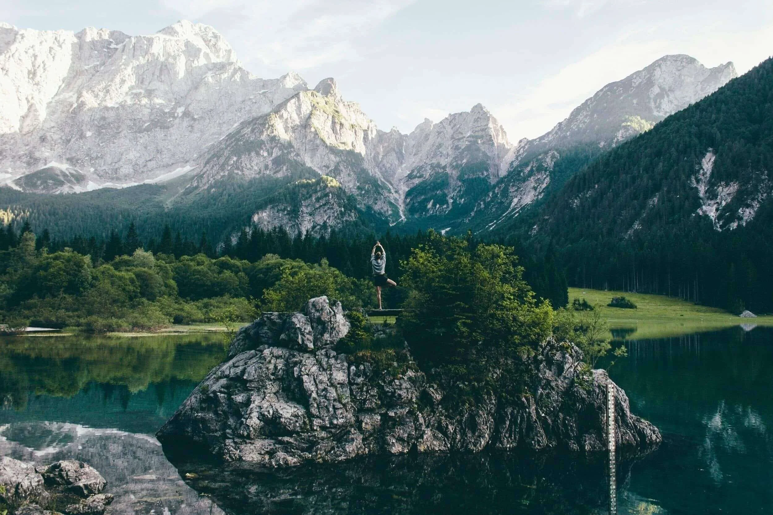 Person doing yoga on a large rock in a serene lake, surrounded by lush greenery and tall mountains in the background.
